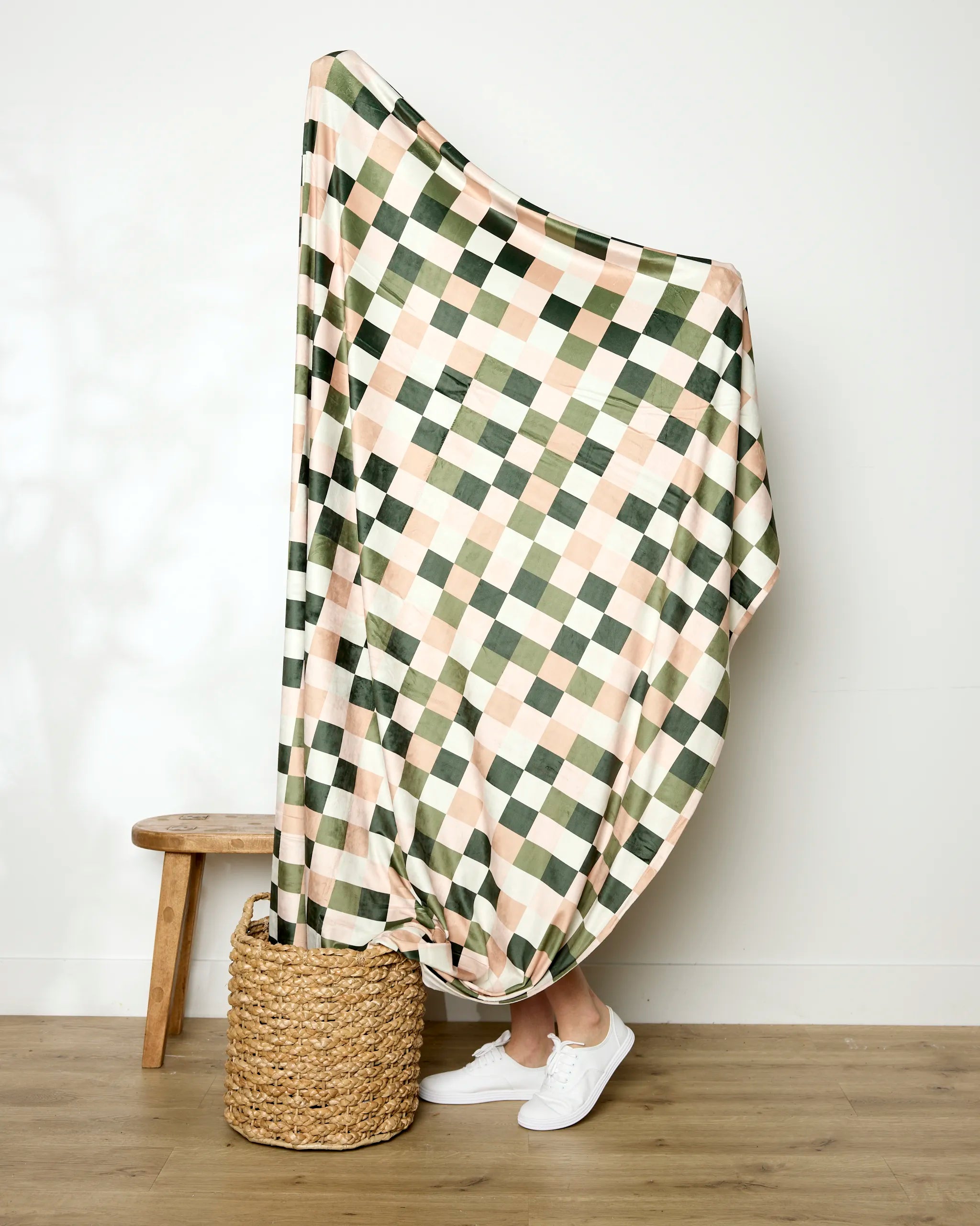 A person stands behind the Geometry Luxora Butter Blanket in green, beige, and pink checks. Only their lower legs and white sneakers are visible as the soft throw drapes over a woven basket and partially covers a wooden stool.