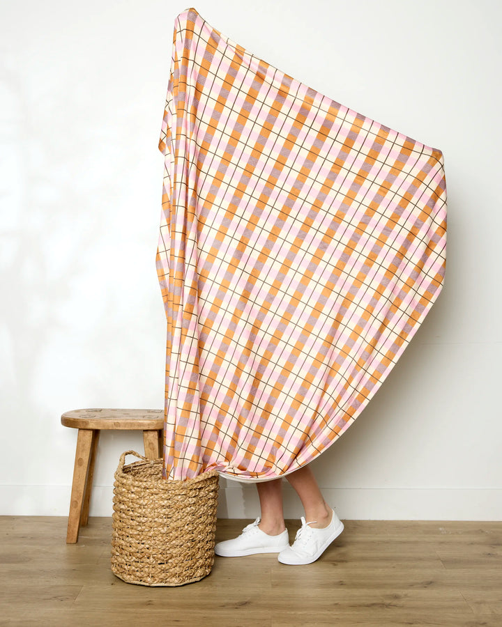 A person stands behind a Megan Roy Maple Plaid blanket, holding it up and covering themselves. Only their legs, white shoes, a wooden stool, and a woven basket are visible against a plain wall.