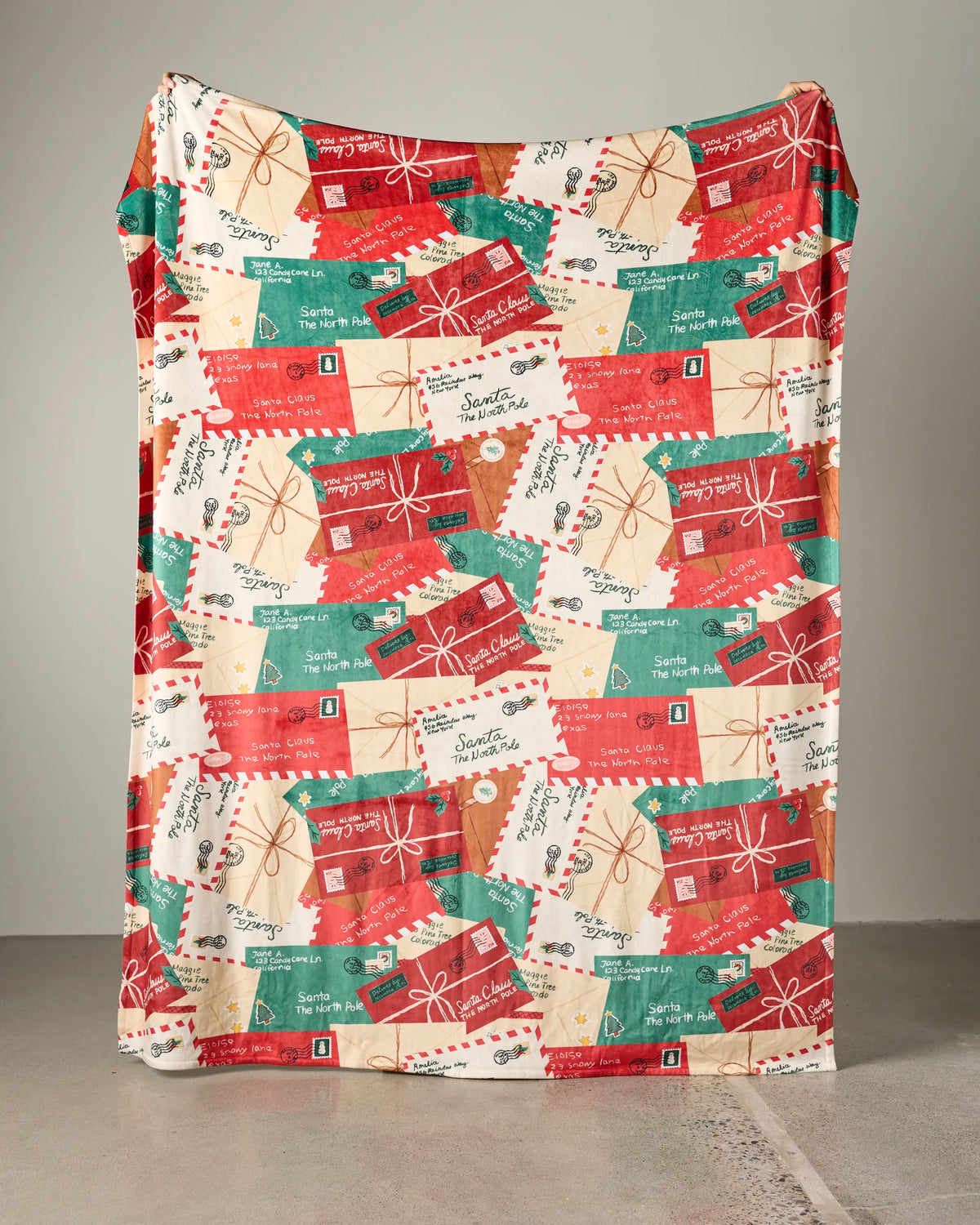 The Geometry Merry Mail throw blanket, featuring a festive pattern of colorful Christmas gifts and postage stamps in red, green, and cream tones, is draped against a plain background.