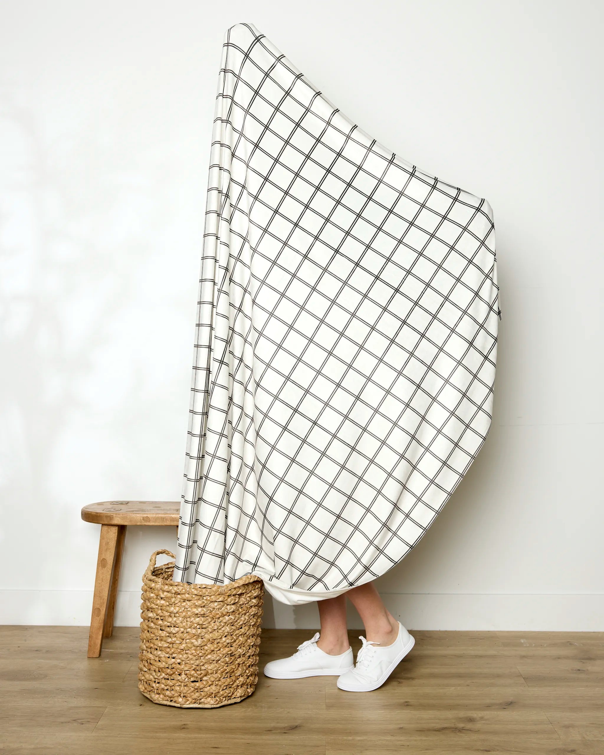 A person stands behind the large white "Moon" throw by Geometry, featuring a black grid pattern and hiding their body. Only their lower legs and white shoes are visible, with a woven basket and small wooden stool nearby.