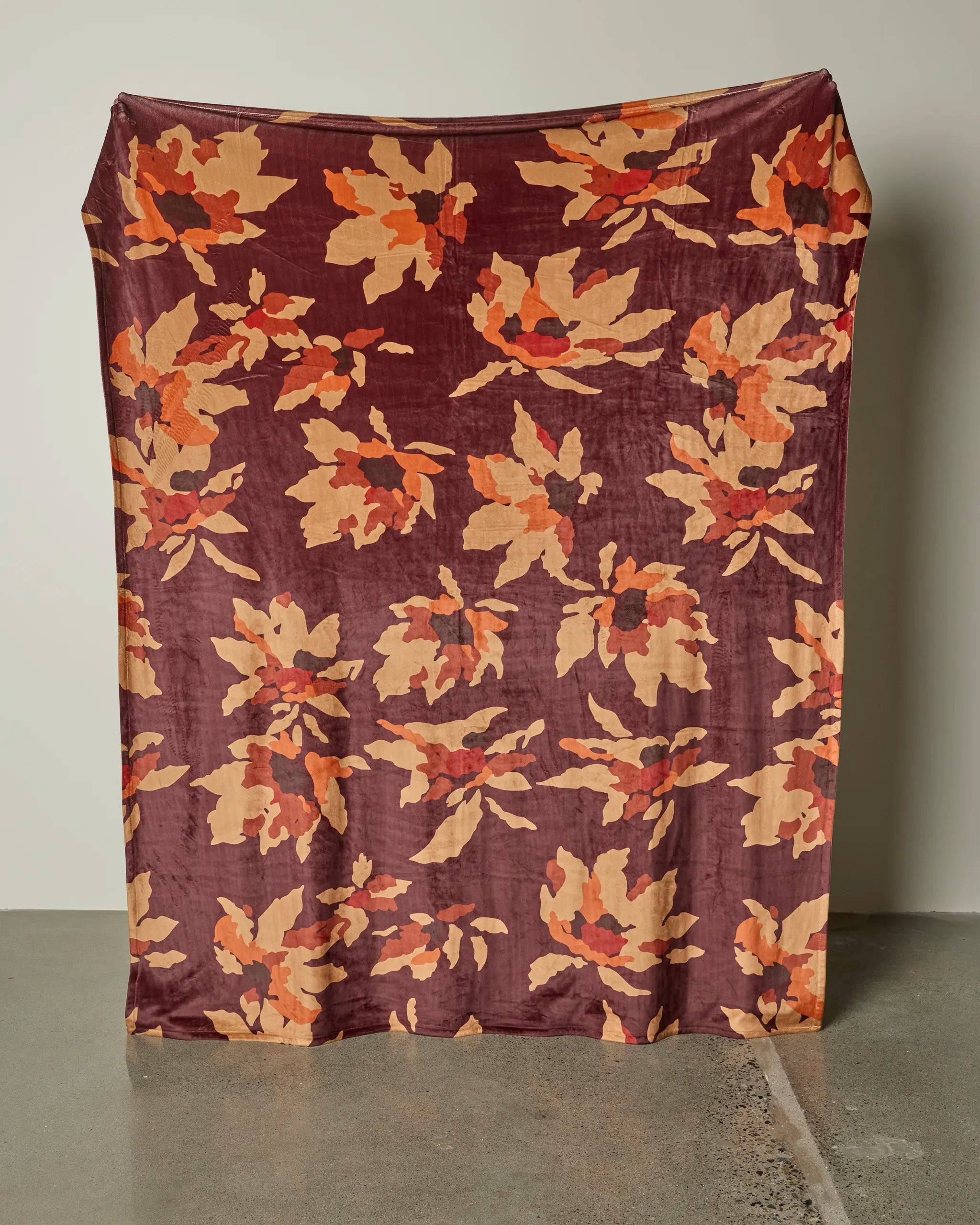 The Mulberry Bloom butter blanket by Geometry, featuring an orange and beige floral pattern on maroon, is draped flat against a white wall and reaches down to the polished concrete floor.