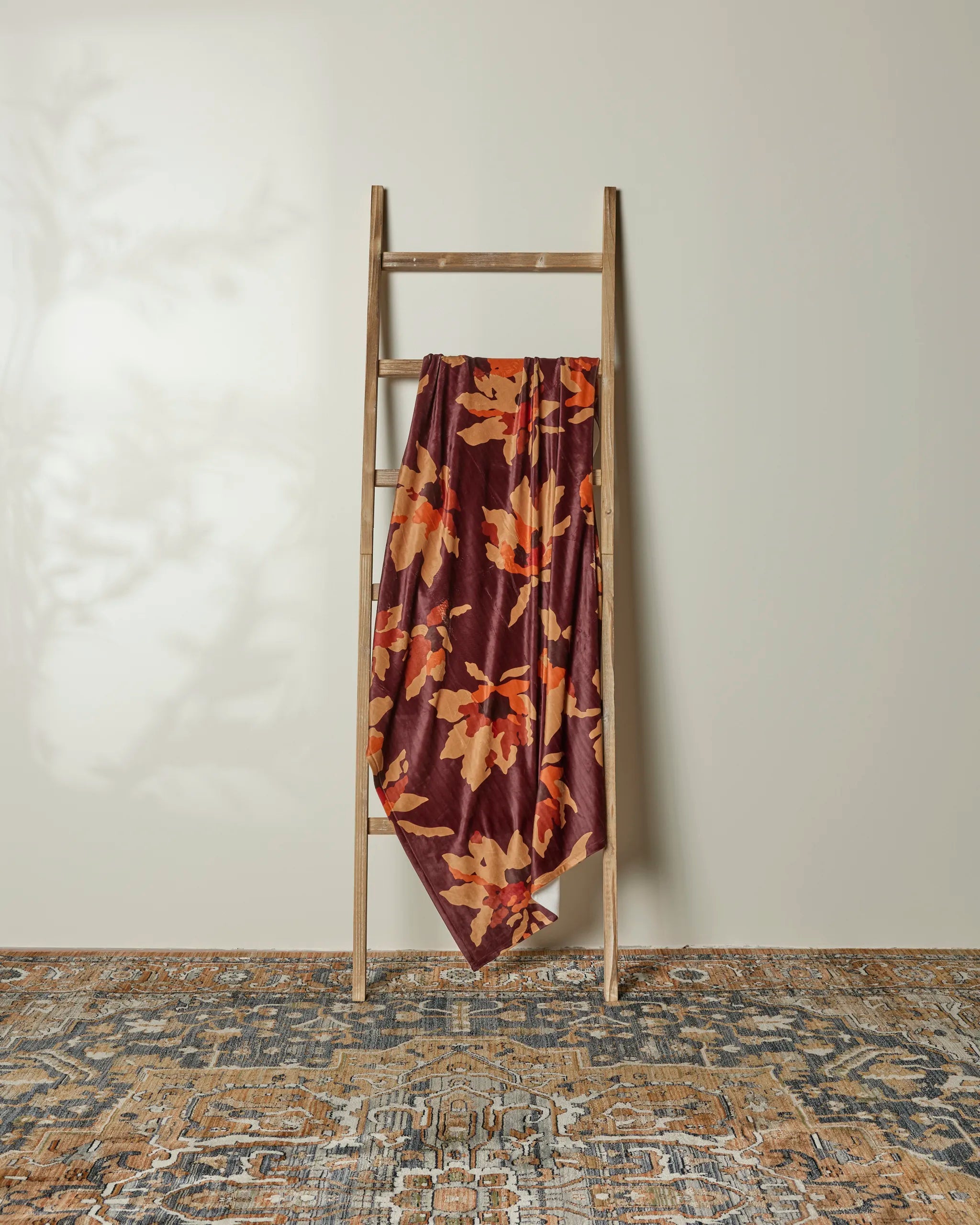 A wooden ladder leans against a light wall, with Geometry’s Mulberry Bloom recycled blanket—featuring a bold orange and yellow floral pattern—draped over one rung. An ornate blue and tan rug lies on the floor.