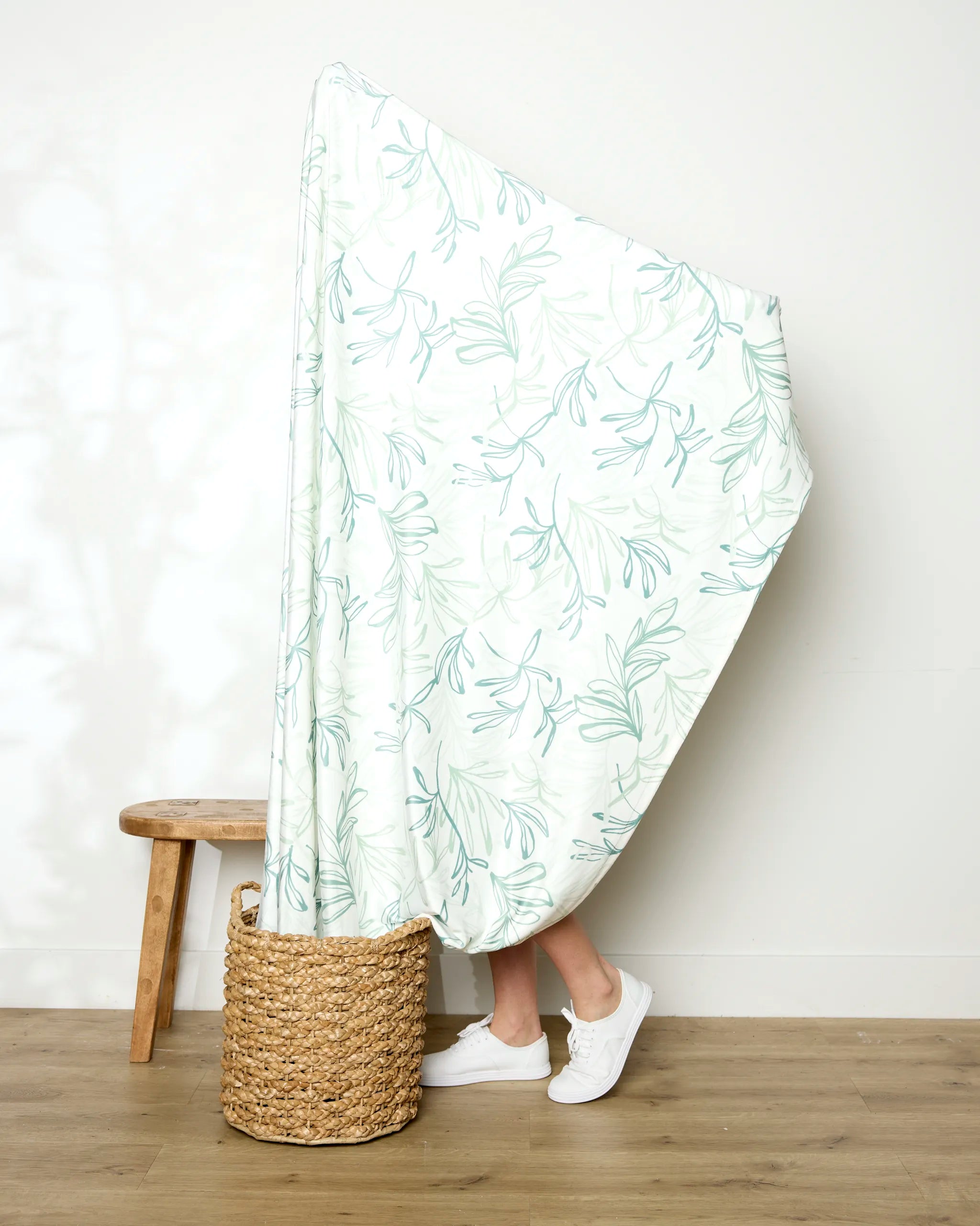 A person stands in a woven basket, mostly covered by the Kris Ruff "Organic Foliage" throw. Only their lower legs and white sneakers are visible. A wooden stool is nearby against a plain white wall and wood floor.
