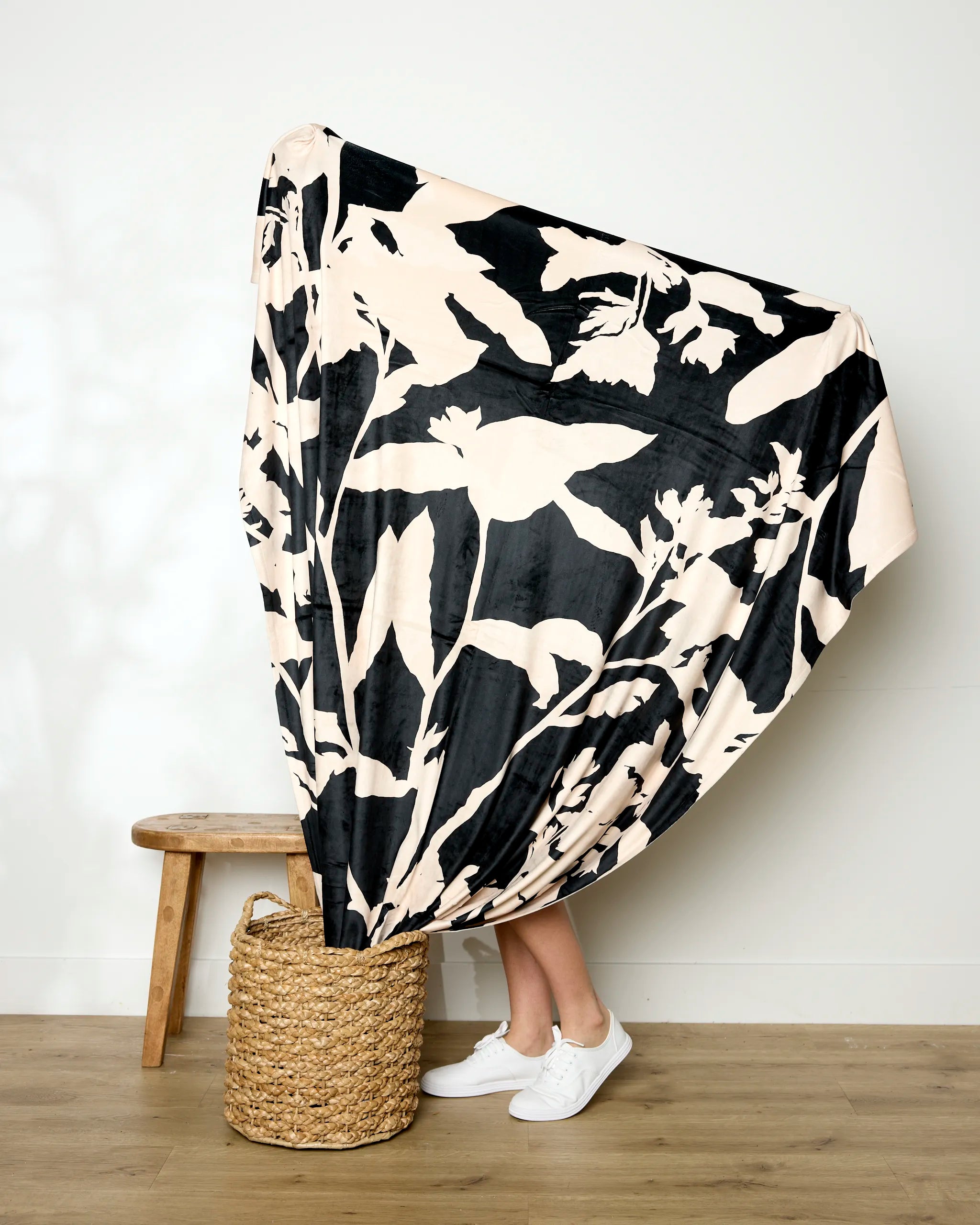 A person stands with legs visible behind a silky soft Pam blanket by Geometry, featuring a black-and-white floral pattern, draped over their upper body beside a wooden stool and woven basket on a light wood floor against a white wall.