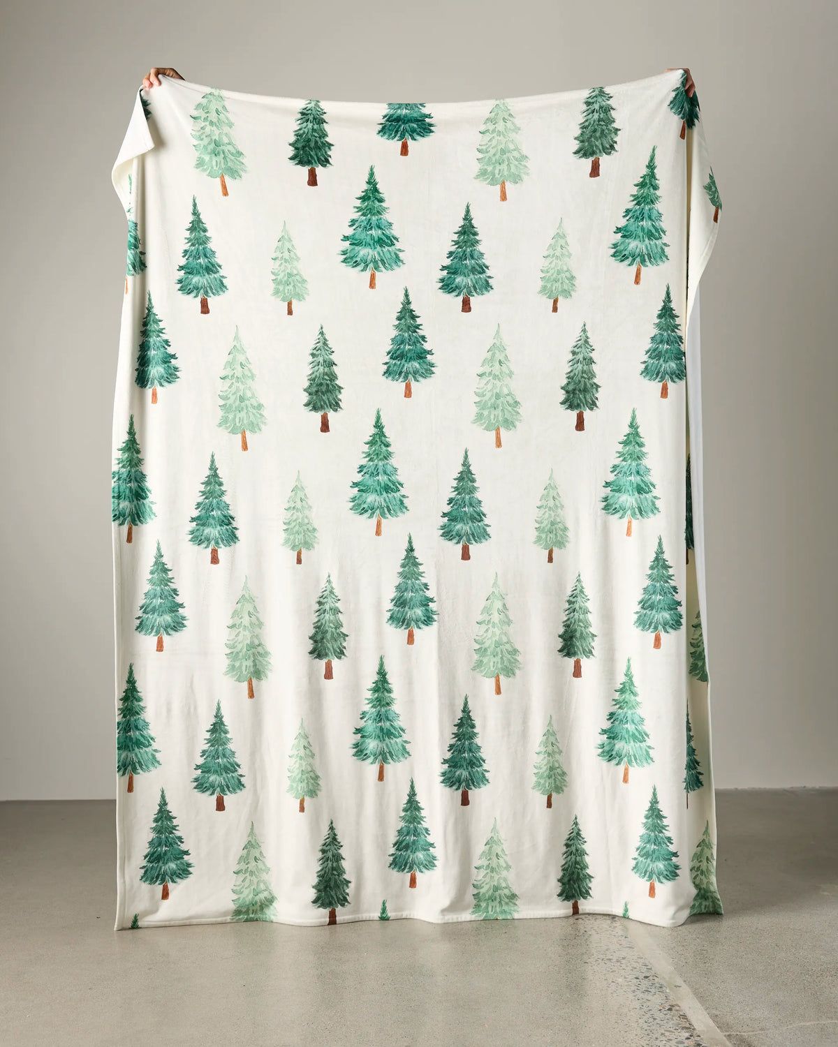 A person holds up Geometry’s Pine Grove Butter Blanket, featuring an all-over pattern of green Christmas trees in various shades on a large white background.