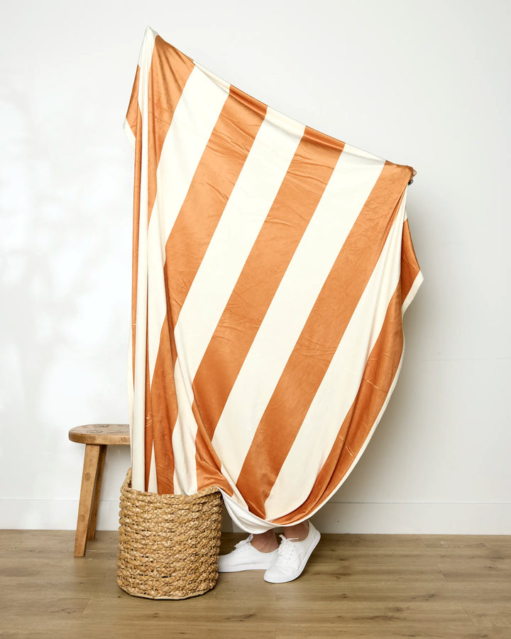 A person stands behind the Geometry Radiance throw blanket, featuring bold orange and white stripes, with only their feet in white sneakers visible beside a woven basket and wooden stool on a wooden floor.