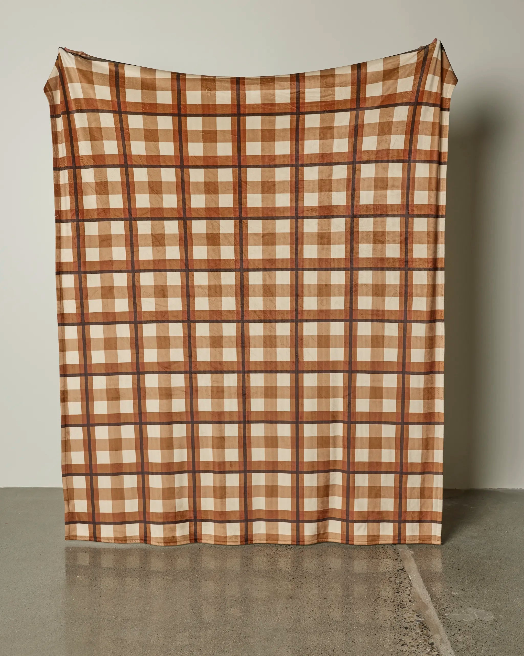 A large Geometry Rich Cocoa blanket in brown, tan, and cream plaid hangs against a plain light wall, draping down to the polished concrete floor.