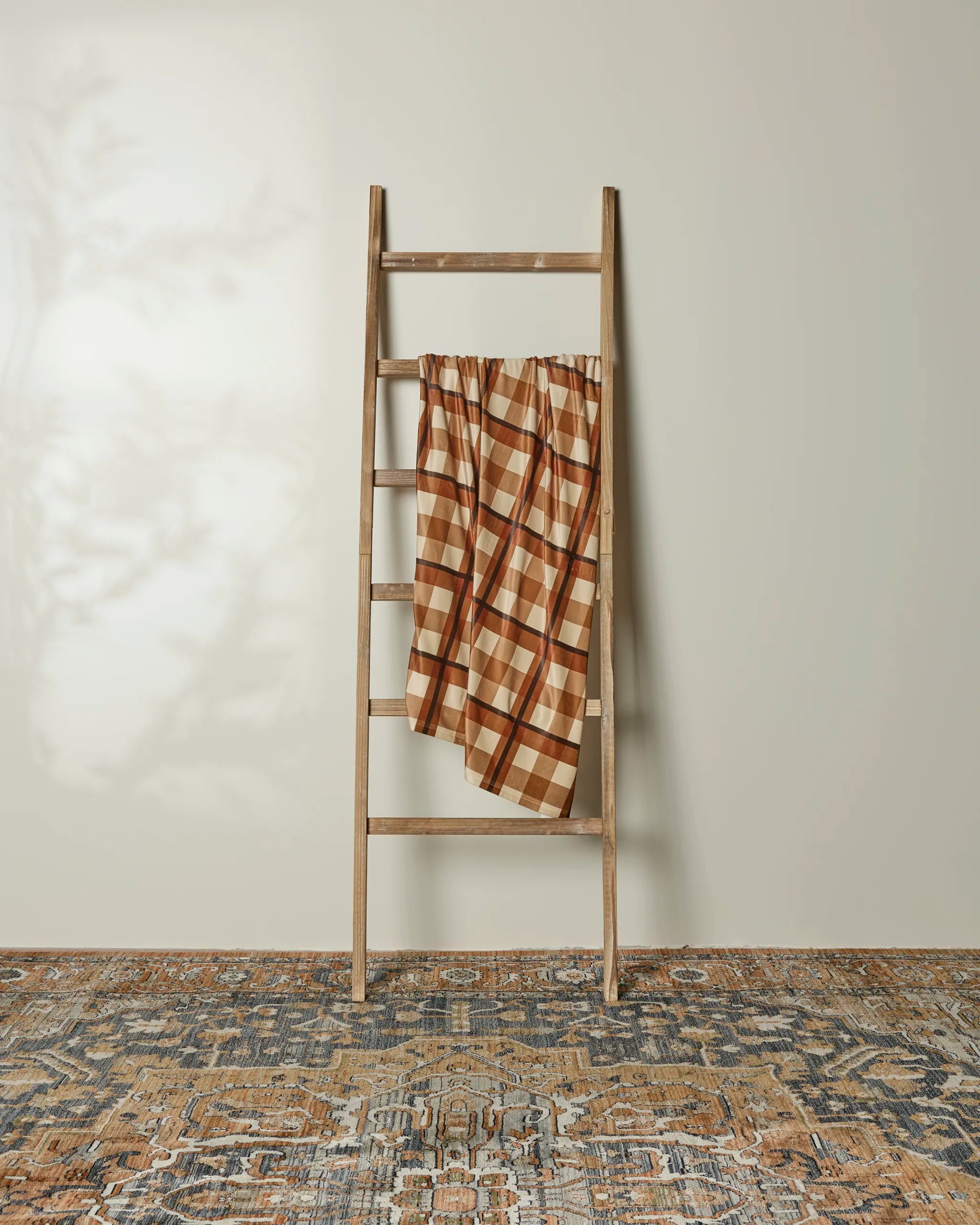 A wooden ladder leans against a beige wall, draped with Geometry’s Rich Cocoa recycled blanket. An intricately patterned rug lies on the floor, while soft natural light and shadows create a calm, minimalist vibe.