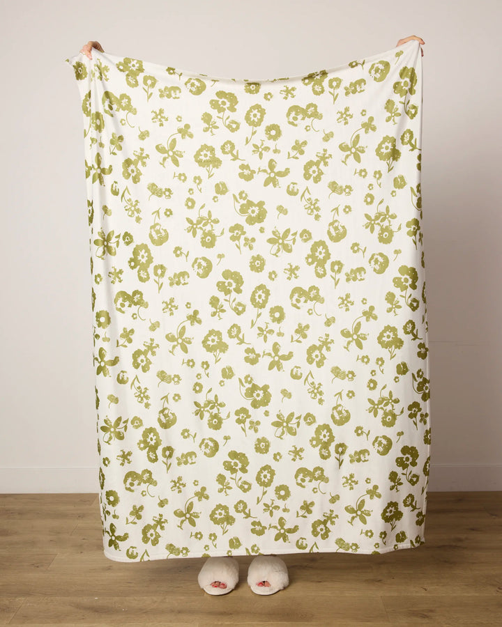 A person stands on a wooden floor, holding up the "Singing Forest" throw blanket by Geometry, featuring a green floral pattern. Their body is wrapped in the blanket, with only their feet in fuzzy white slippers showing against a plain white wall.