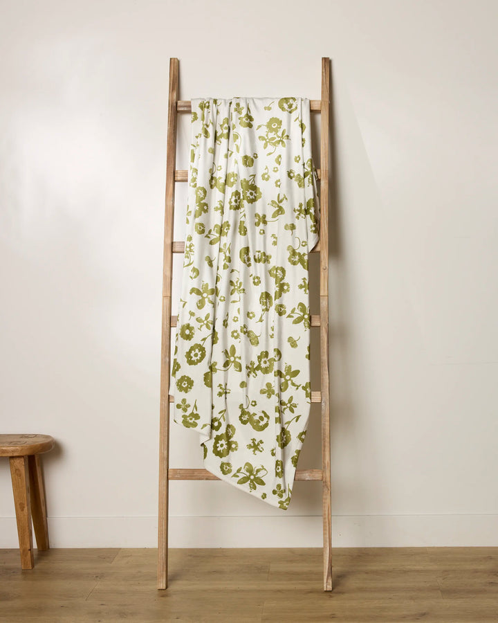 A wooden ladder leans against a beige wall with Geometry’s Singing Forest—a soft throw blanket featuring a green floral pattern—draped over it; a small wooden stool sits to the left.
