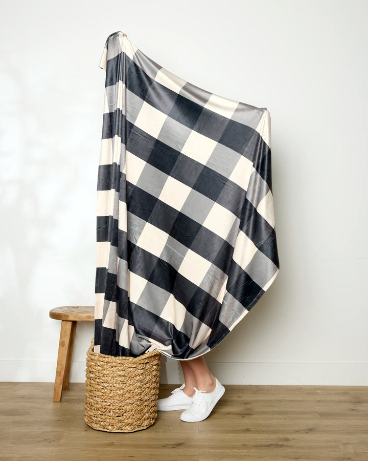 Wrapped in Geometry’s Snowy Gingham recycled blanket, a person stands in a woven basket by a wooden stool, showing only their lower legs and white sneakers in a minimal room.