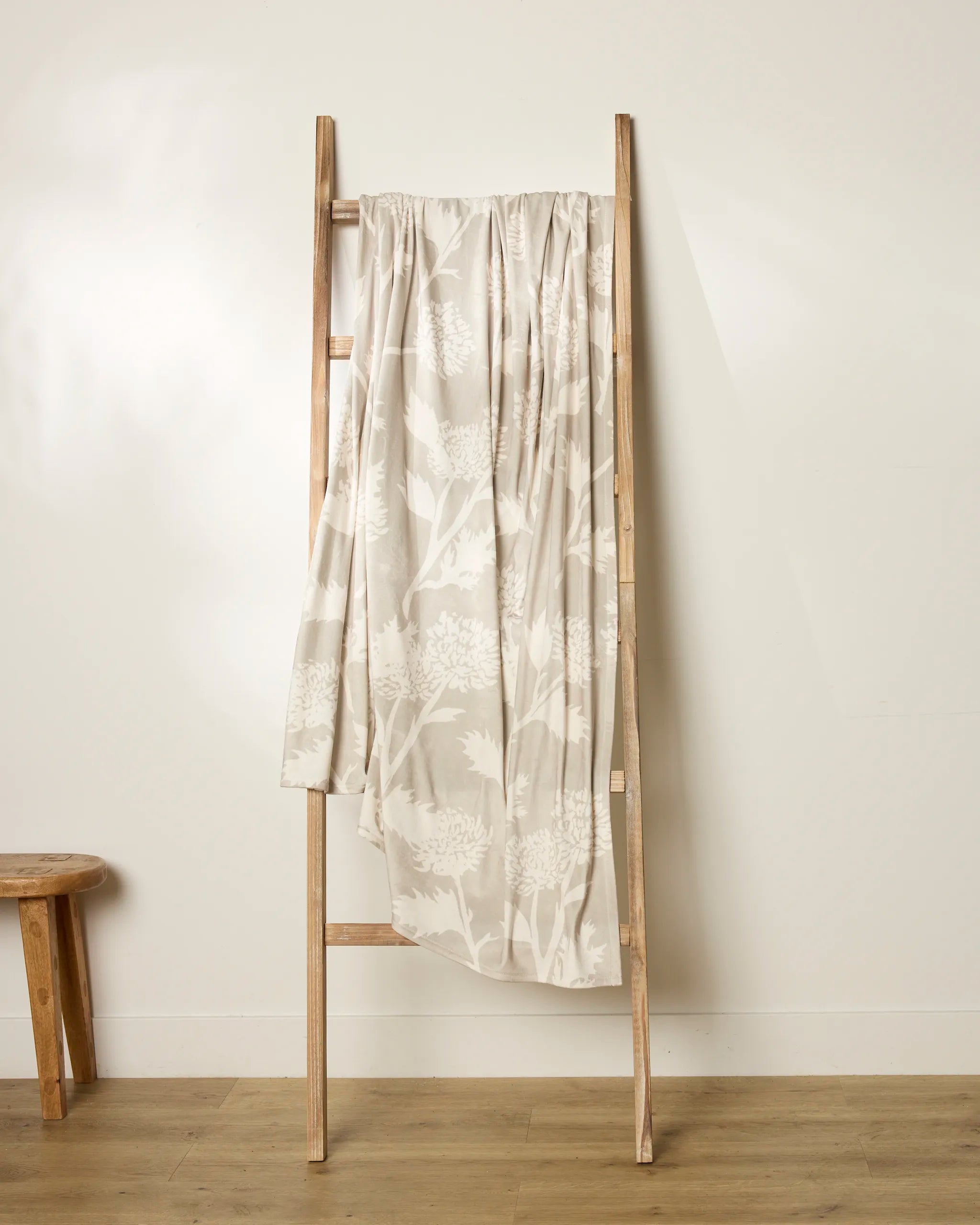 A light wooden ladder leans against a white wall, draped with Geometry's Soft Blooming Thistle—a cream-colored, floral-patterned throw blanket. A small wooden stool sits to the left on a light wood floor.