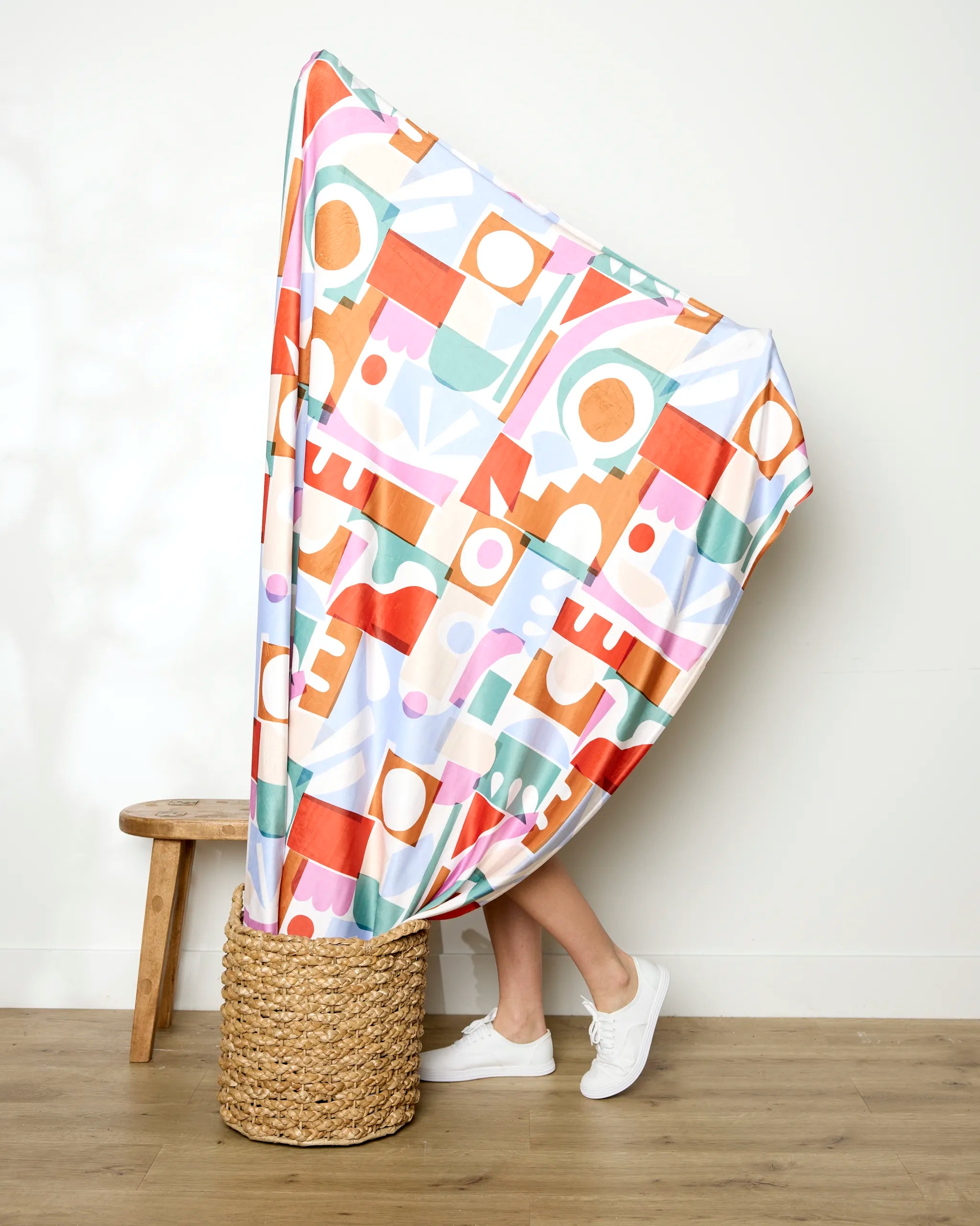 A person in white shoes stands in a woven basket, mostly covered by Claire Iglesias’ Soft Cutouts Garden blanket with colorful geometric patterns, beside a wooden stool against a plain light wall.