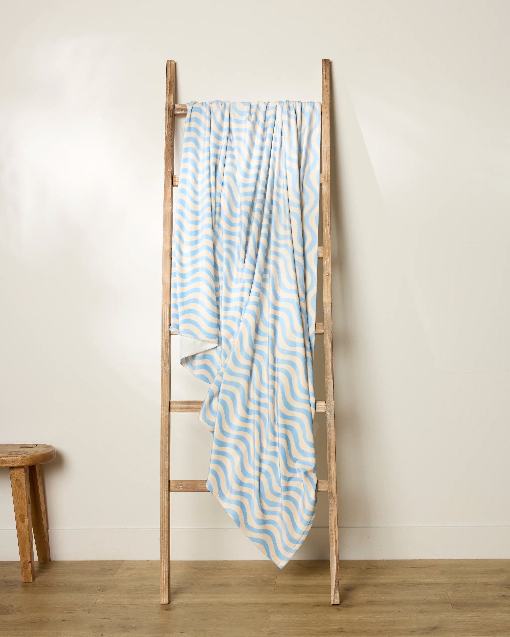 The Geometry Spatial ladder, in light wood, leans against a white wall with a blue and yellow wavy striped throw draped over it; a small wooden stool sits on the wooden floor to the left.