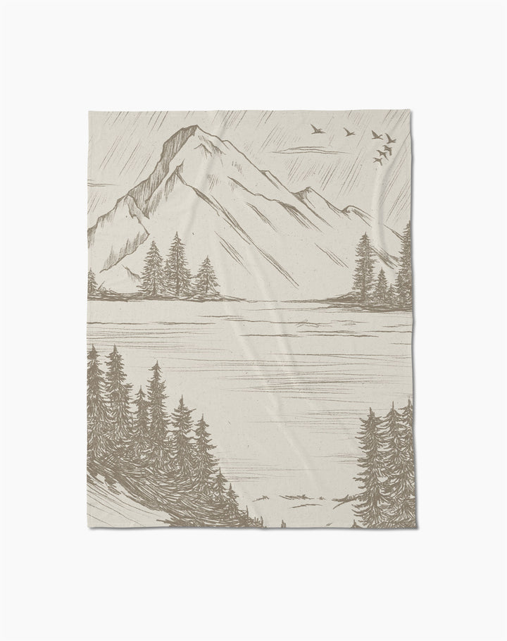 The Geometry Stillpine throw blanket, in textured beige, displays a detailed mountain landscape with pine trees, a lake, and birds in flight.