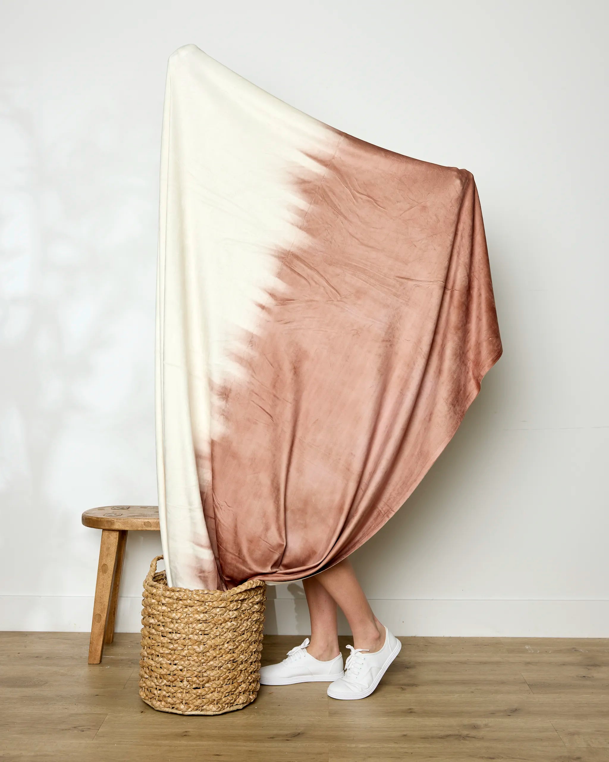A person holds up the large Geometry Sundae Butter Blanket, featuring a white and blush pink tie-dye pattern. Only their lower legs and white sneakers are visible, with a woven basket and wooden stool nearby, showcasing its silky softness.