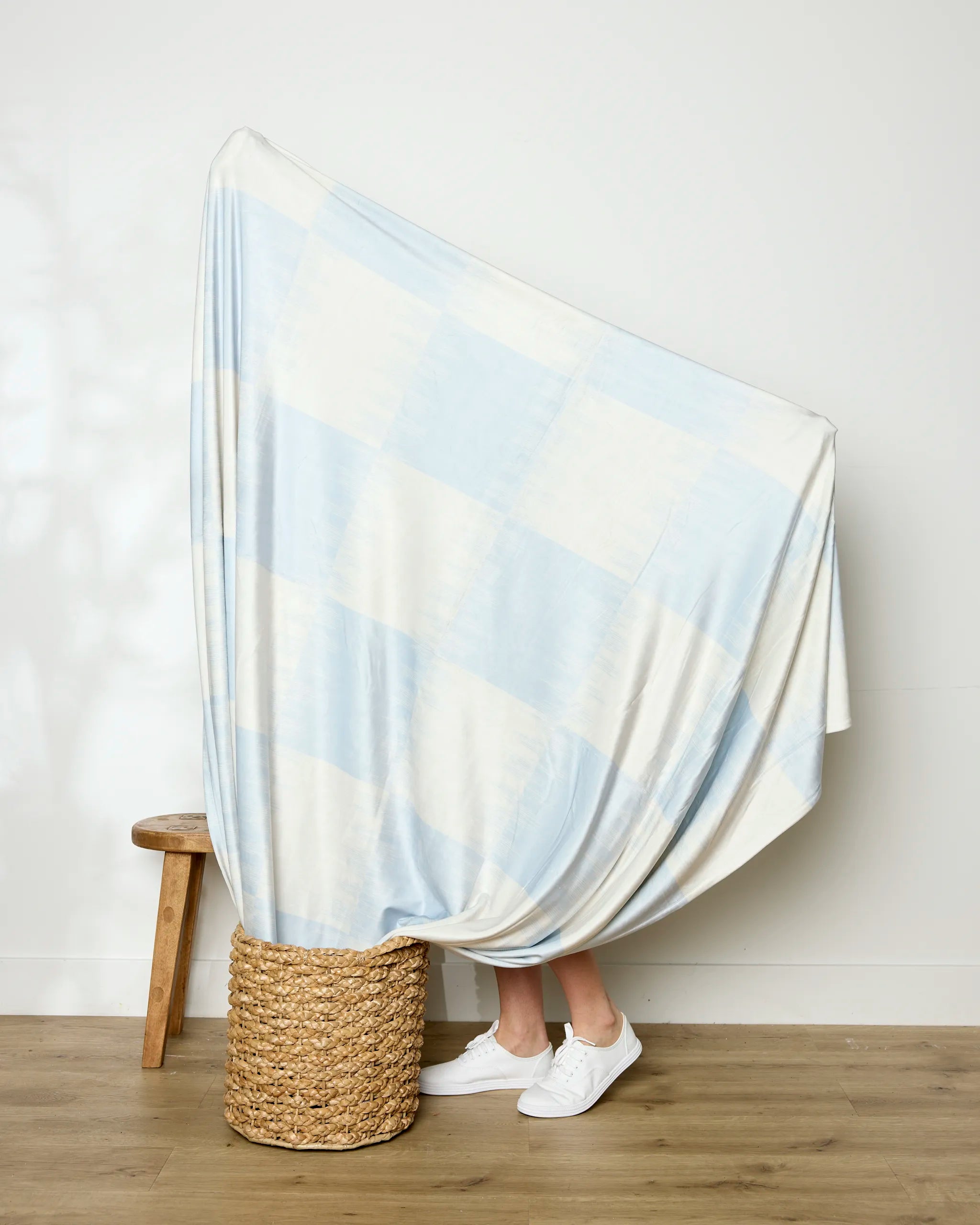 A person stands behind Geometry's Sweet Checks throw blanket in light blue and white, draped over a wicker basket; only their legs and white sneakers are visible beside a wooden stool.