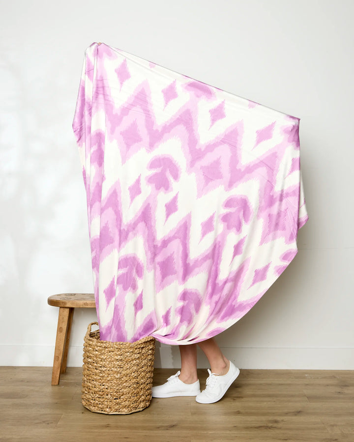A person stands behind the Geometry Tulip Scatter recycled blanket, showing only their legs and white sneakers. The white and purple patterned blanket drapes above a woven basket next to a wooden stool on a wooden floor.