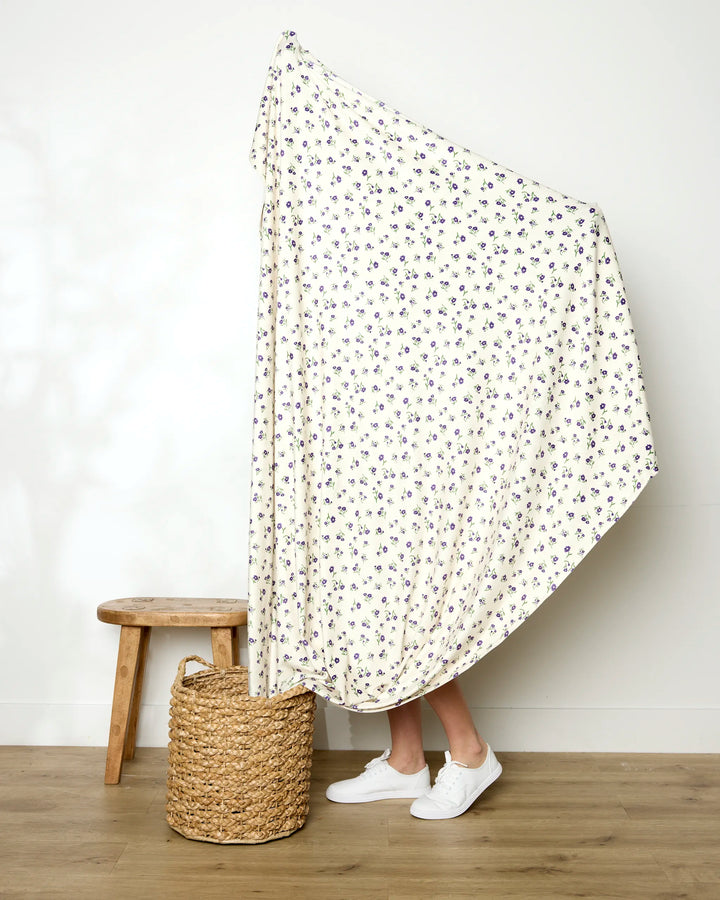 A person stands behind a large Geometry Violet Sound throw blanket, mostly hidden except for their lower legs and white sneakers. The soft blanket drapes over a woven basket beside a wooden stool on a wooden floor.