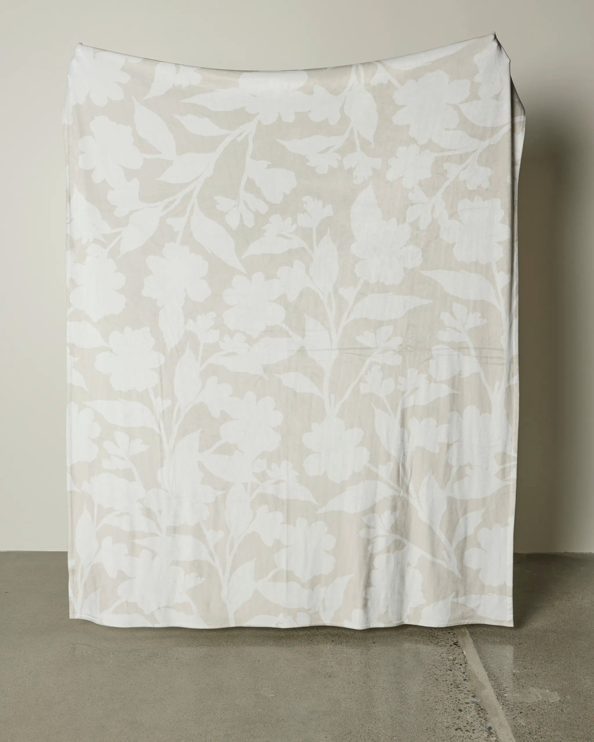 The Whispering Petals by Geometry, a large semi-sheer white fabric with a subtle floral design, drapes elegantly against a light wall and gently touches the smooth concrete floor.