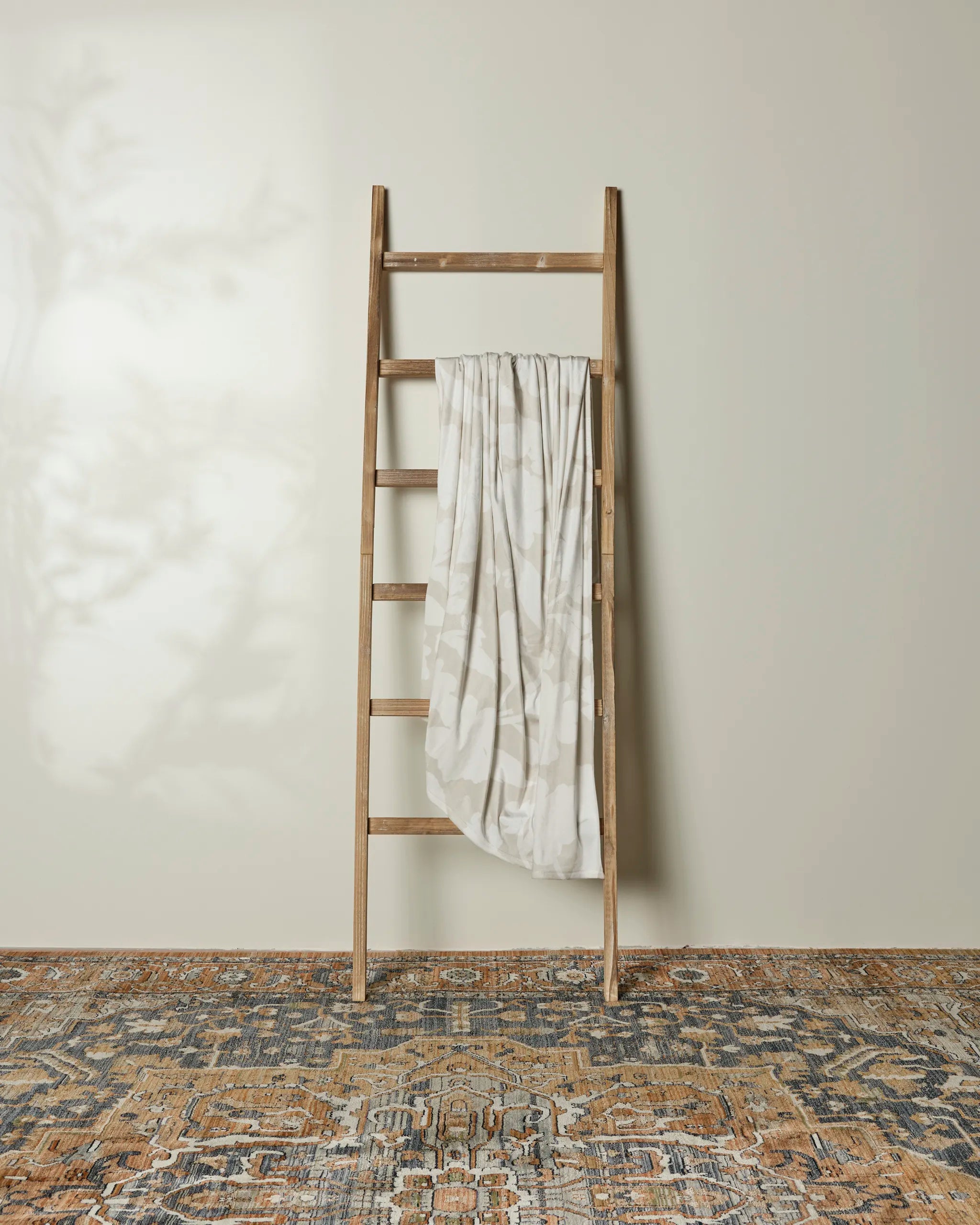 A Whispering Petals blanket by Geometry is draped over a wooden ladder against a light wall. The floor below features an earthy patterned rug, with subtle foliage shadows softening the scene.