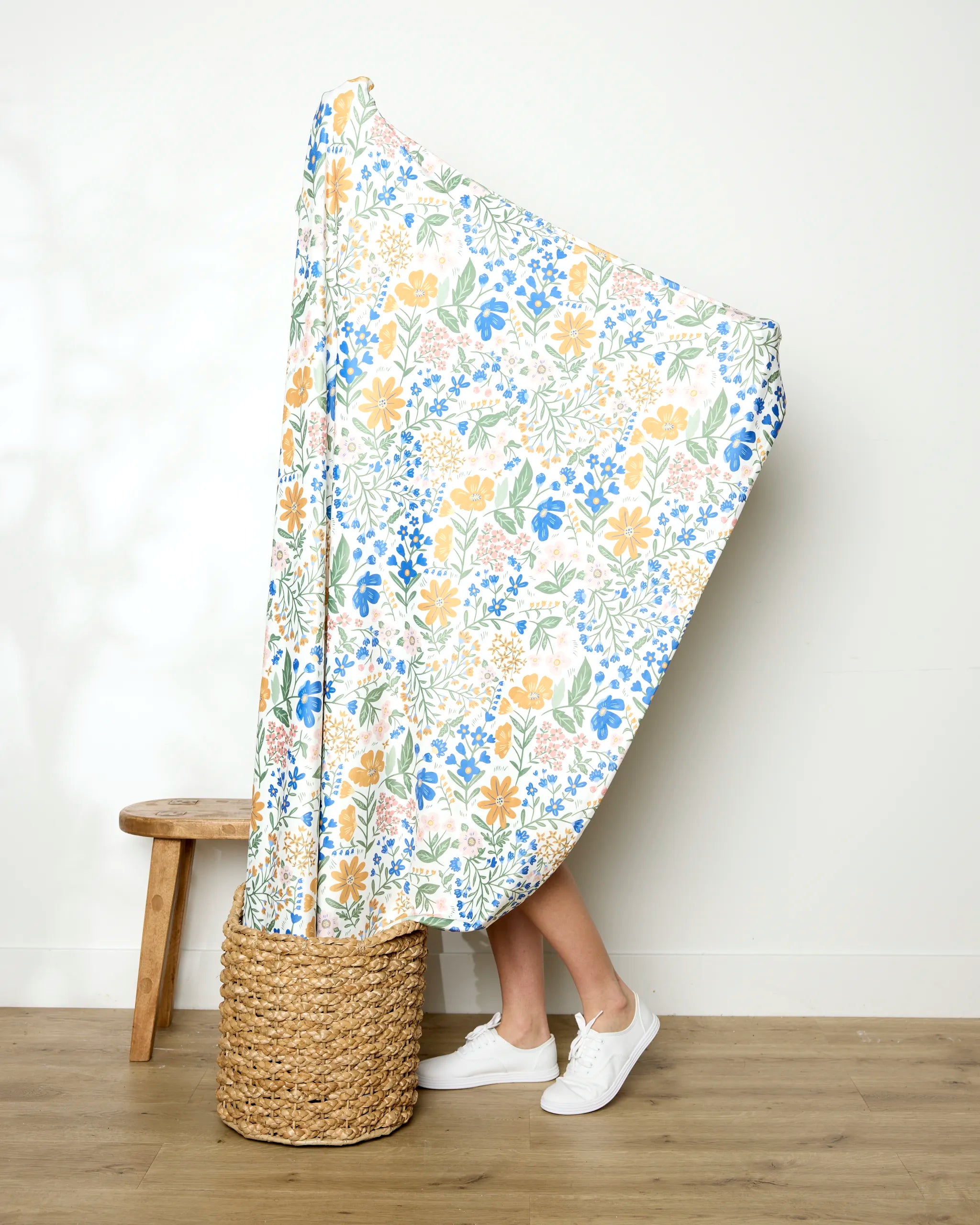 A person stands in a woven basket, mostly covered by the colorful Wildflower Vine Blush blanket from Danelys Sidron. Only their lower legs and white sneakers show. To the left, a wooden stool rests against a plain wall.