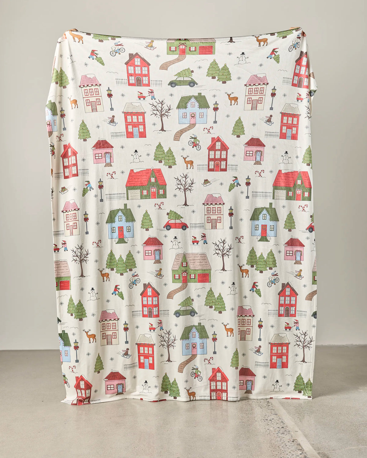 A large piece of fabric from Geometry, called “Winter Village,” reminiscent of a soft throw blanket and featuring a colorful print of houses, trees, cars, people, and animals, is draped vertically against a plain wall, resting on a concrete floor.