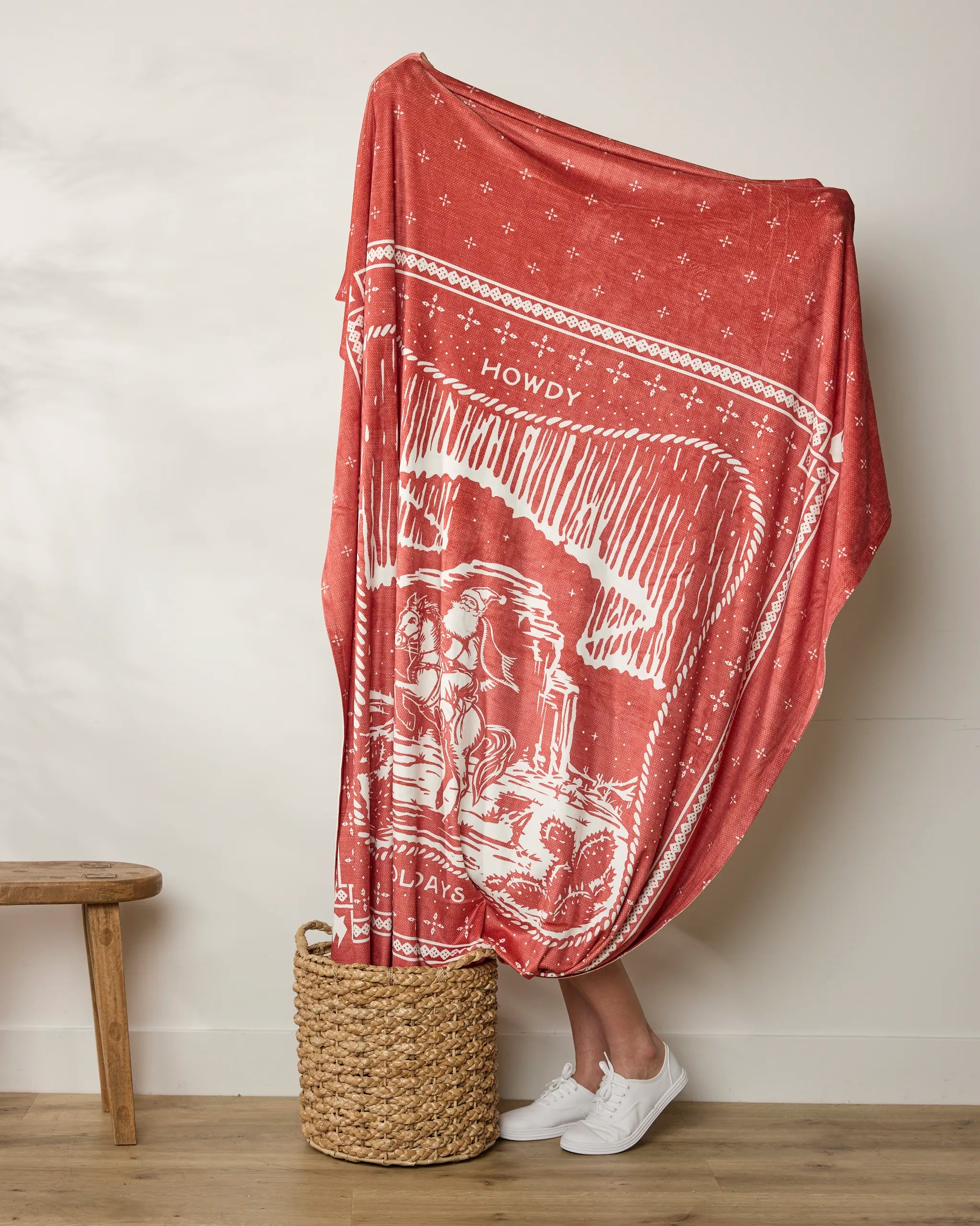 A person stands behind a large red patterned Geometry "Cowboy Christmas" Butter Blanket with white designs and "HOWDY" visible. Only their lower legs in white sneakers show. The soft throw drapes into a woven basket on a wooden floor.
