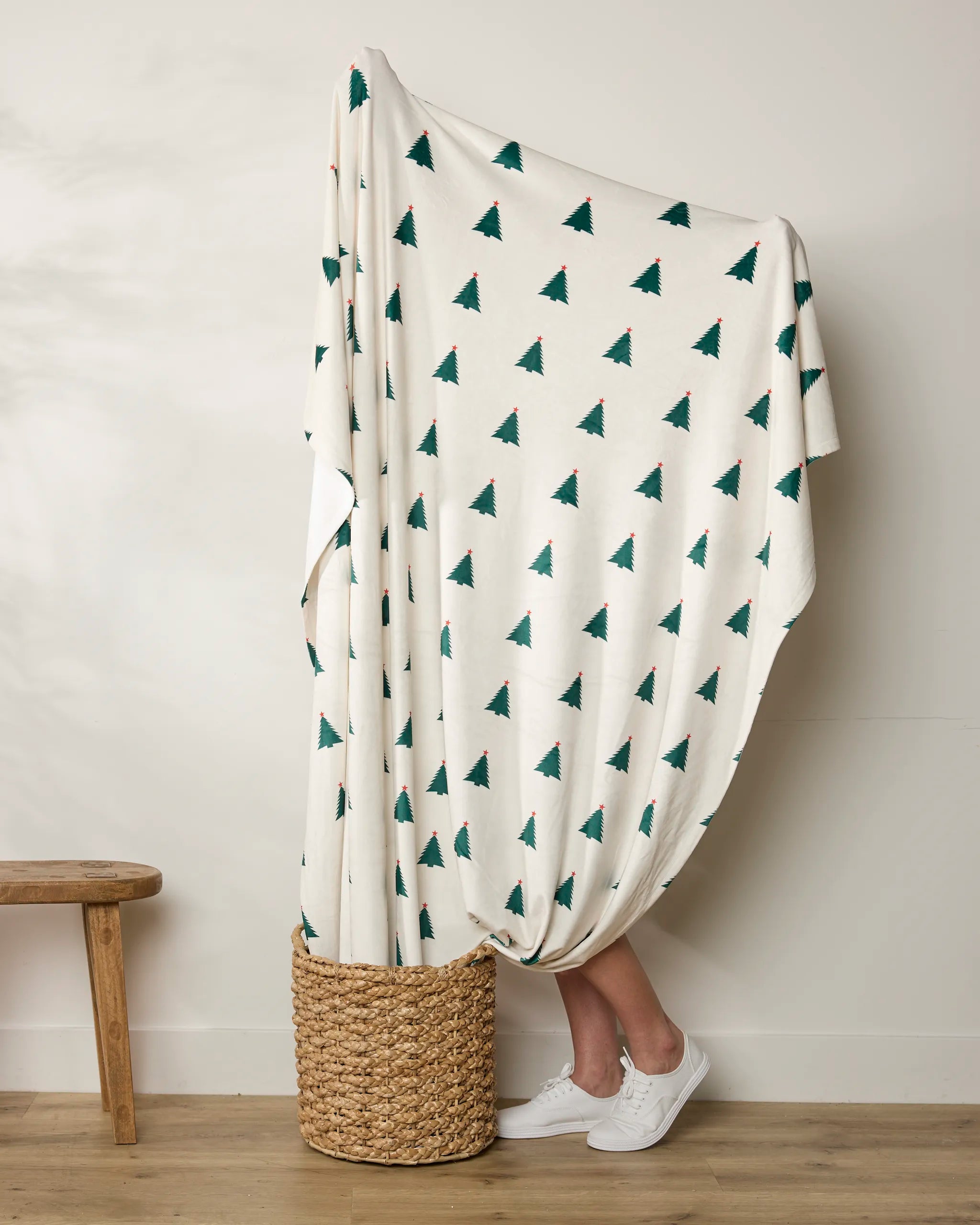 A person stands in a woven basket wrapped in the Geometry Cozy Christmas blanket, featuring green pine tree patterns—only their lower legs and white sneakers are visible. To the left, a wooden bench rests against a plain wall.