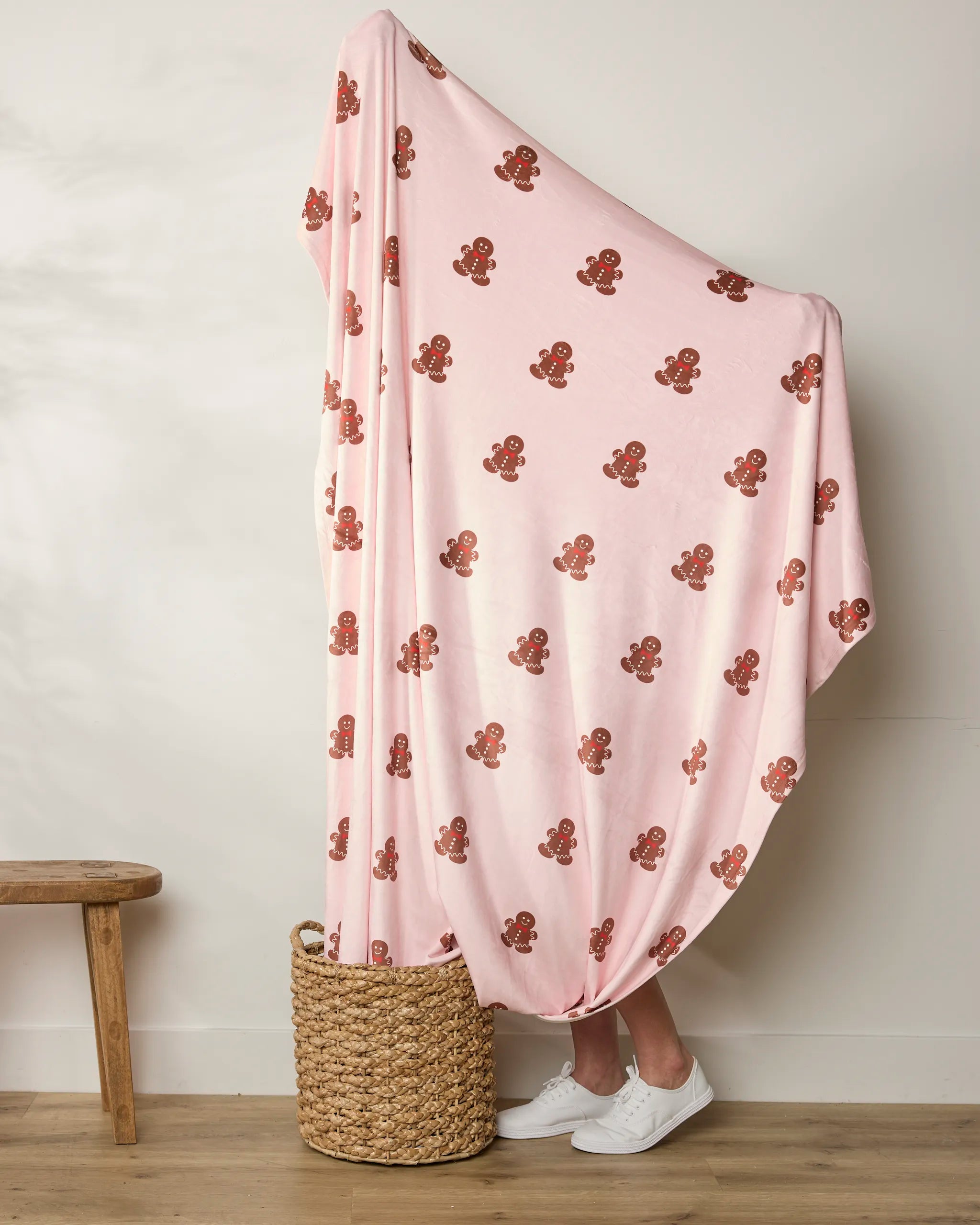 A person stands in a basket, wrapped in Geometry's Cozy Gingerbread throw—a pink, gingerbread men blanket. Only their white sneakers and lower legs are visible beside a wooden bench against a plain wall.