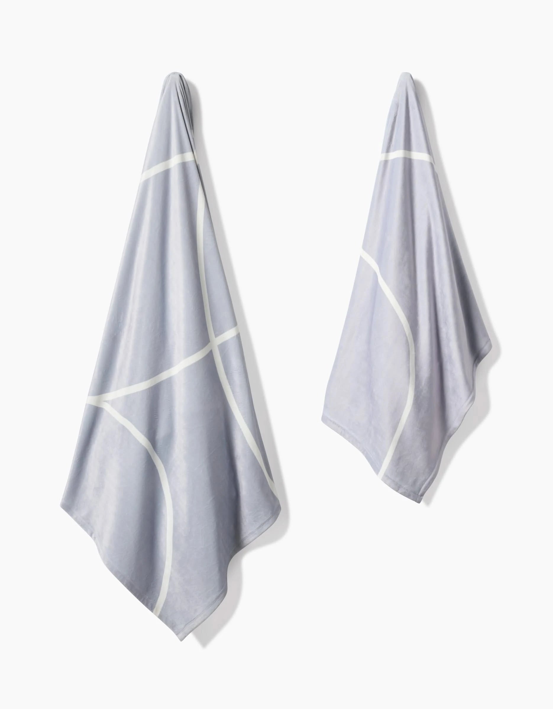 Two light gray Geometry Crossover towels with white curved lines hang side by side on a white wall, evoking the softness and modern, minimalist design of an eco-friendly blanket made from recycled materials.
