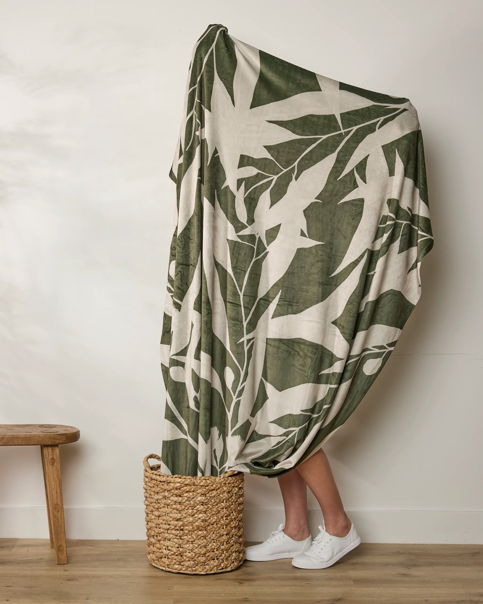 A person stands in a woven basket, mostly wrapped in Geometry's Fable blanket, with only their lower legs and white sneakers showing. A wooden bench sits to the left against a plain wall.
