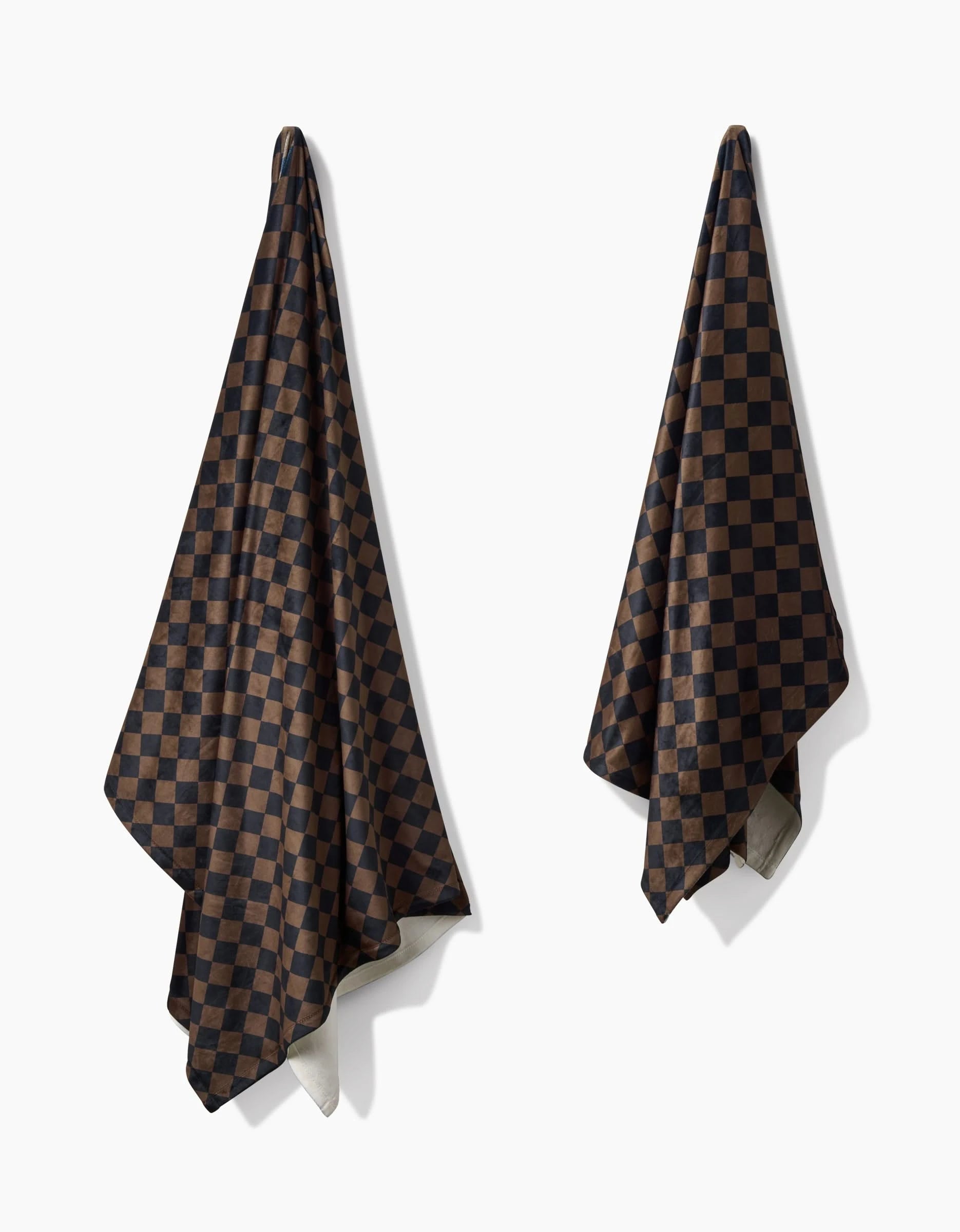 Two Geometry Gridlock towels, featuring a black and dark brown checkerboard pattern, hang separately on a white wall in loose folds, evoking the cozy allure of an eco-friendly blanket.