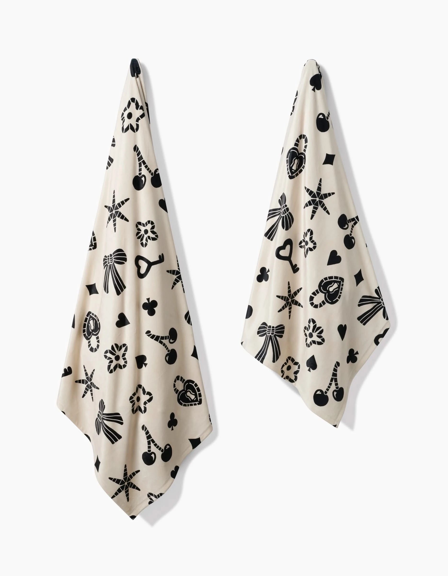 Two beige Heart Locket towels by Geometry, featuring black graphics like bows, clubs, stars, hearts, and locks, hang side by side against a white background. Their silky softness is inspired by the Butter Blanket collection.