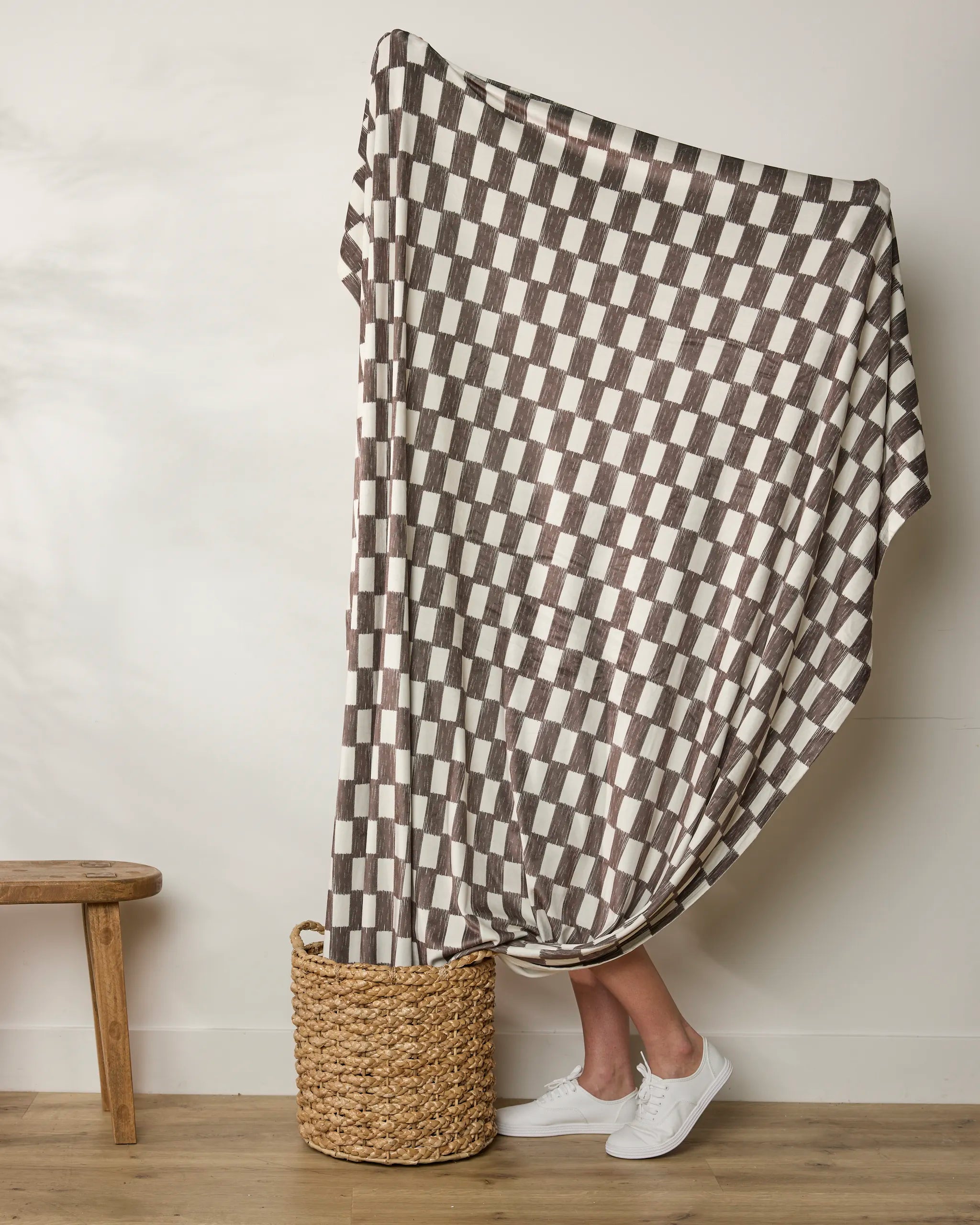 A person stands in a woven basket, mostly covered by Geometry's Hot Coa Coa throw, featuring brown and white checks. Only their white sneakers and lower legs show. A small wooden bench sits to the left.