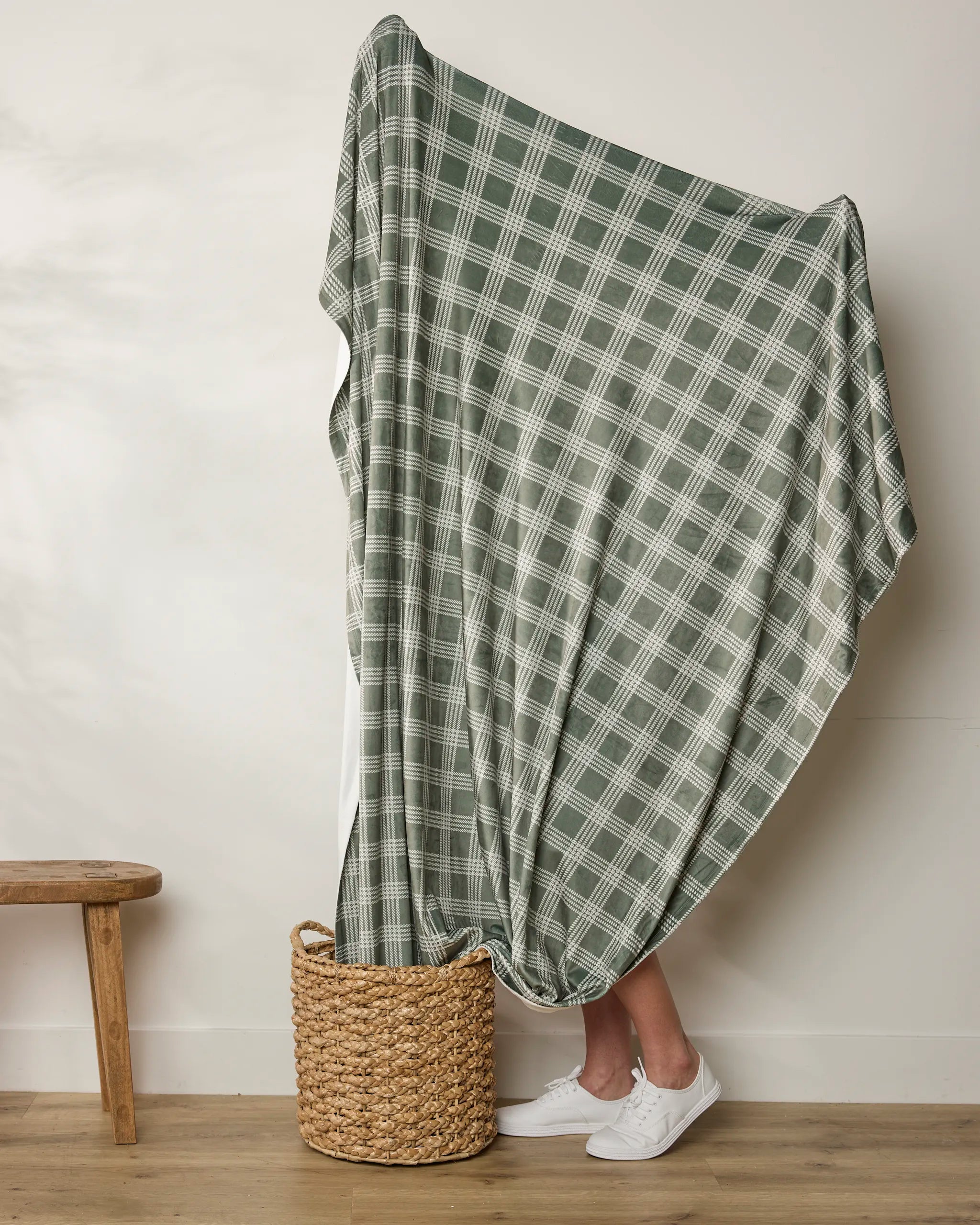 A person stands behind the Geometry Juniper green and white plaid recycled blanket, mostly hidden except for their lower legs and white shoes, with the soft throw draped into a woven basket beside a wooden bench.