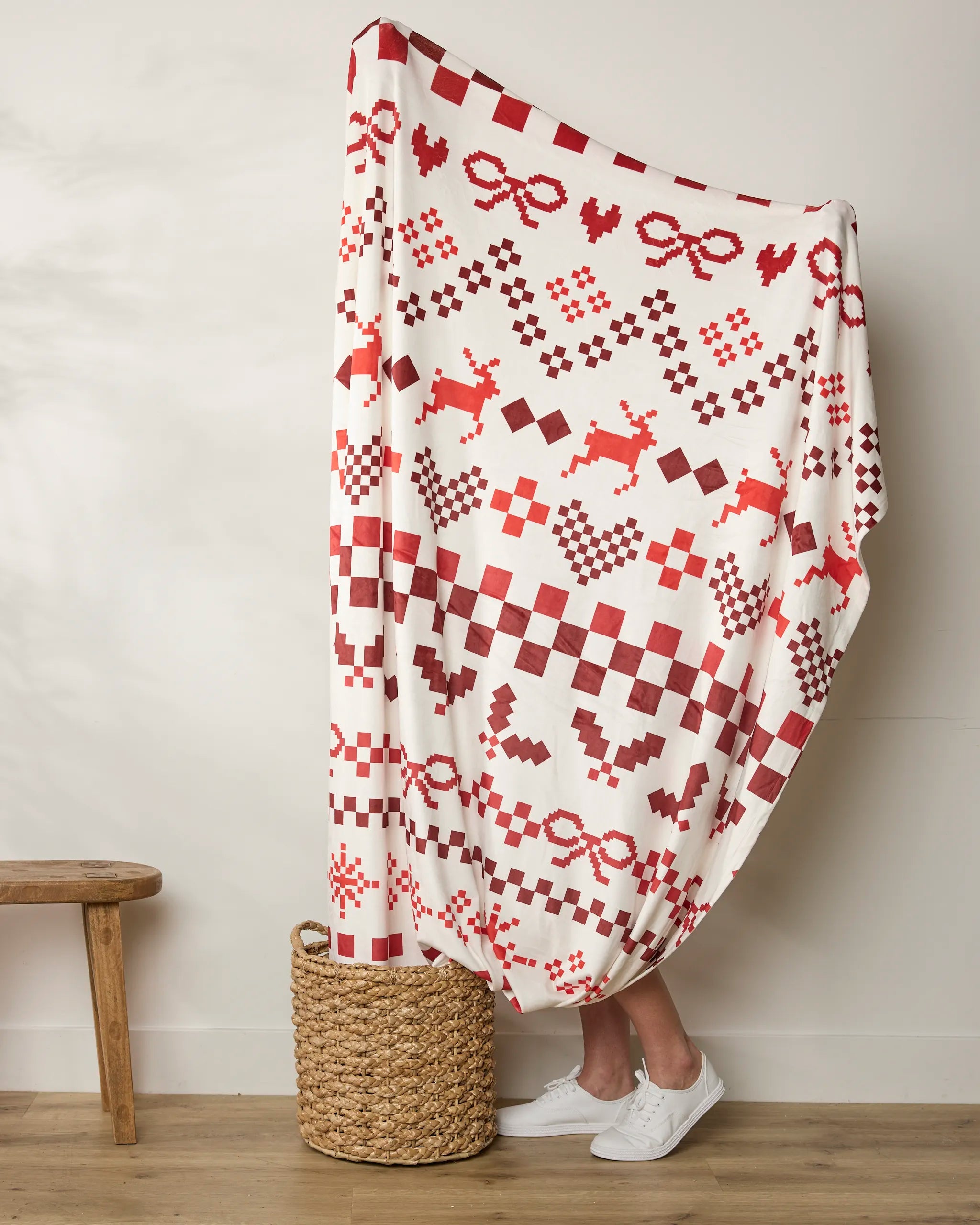 A person stands behind a large white Geometry Merry Pixels blanket featuring red geometric and winter-themed designs like hearts, bows, and reindeer. Only their legs and white shoes are visible. A woven basket and a small wooden bench are nearby.
