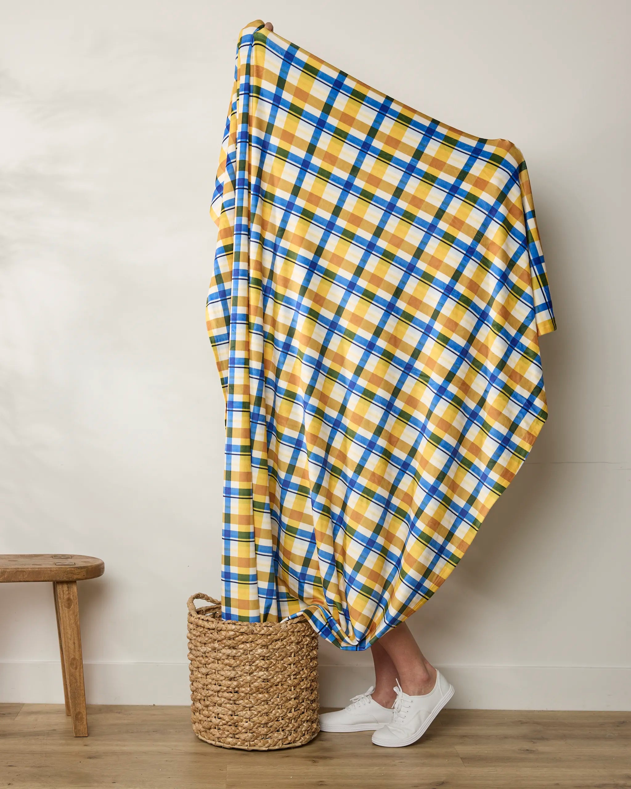 Wrapped almost entirely in the Geometry Nochut throw blanket—featuring yellow, blue, and white plaid—a person stands in a woven basket, with only their lower legs and white sneakers showing; a bench sits to the left against a plain wall.