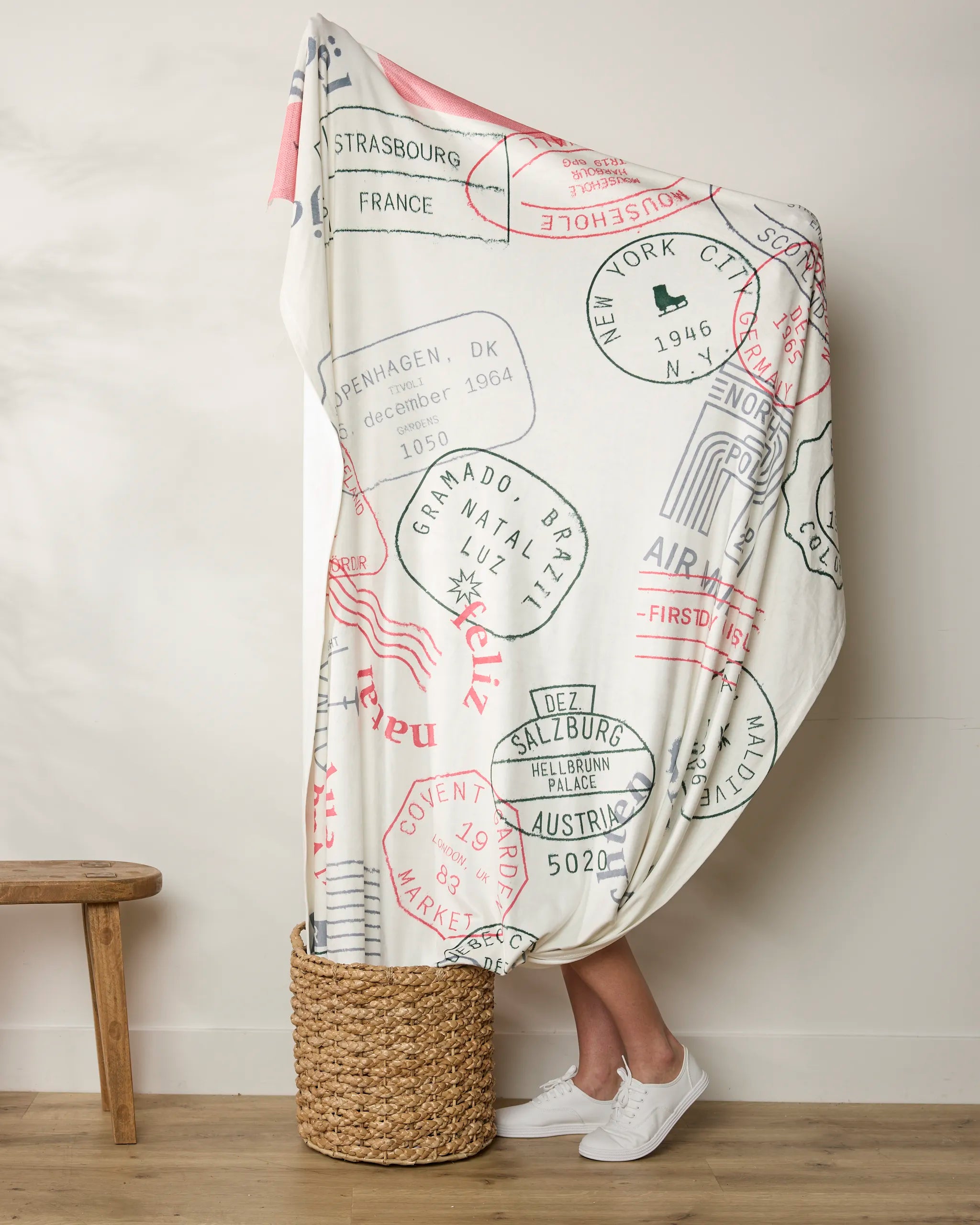 A person stands in a wicker basket, mostly wrapped in the Geometry Nordic Mail Butter Blanket with passport stamp designs; only their lower legs and white sneakers are visible. A plain wall and part of a wooden bench form the background.