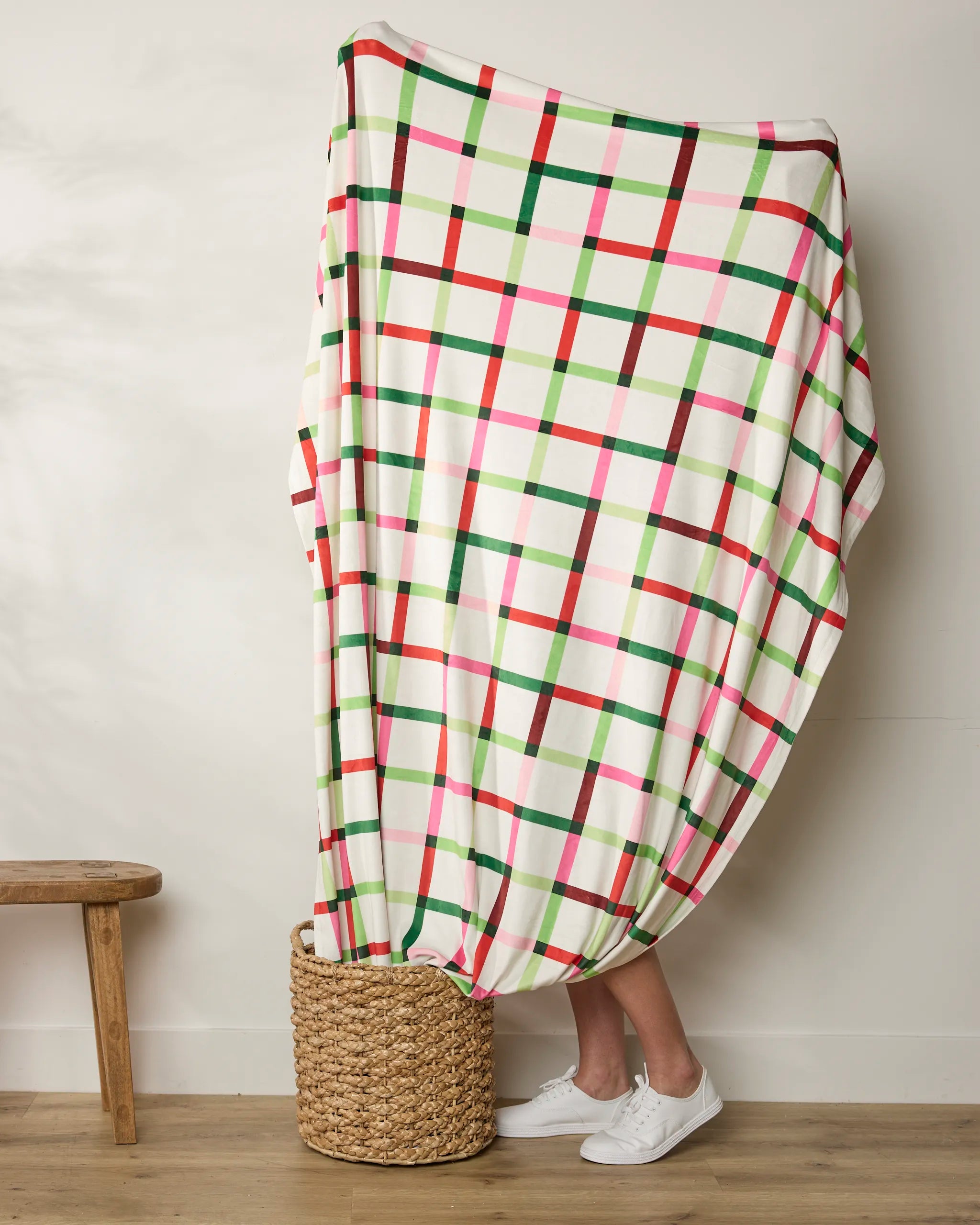 A person stands in a woven basket, mostly draped in Geometry’s Season of Joy blanket—a white recycled throw with colorful plaid stripes—revealing just their lower legs and white sneakers. A wooden bench sits nearby against a plain wall.