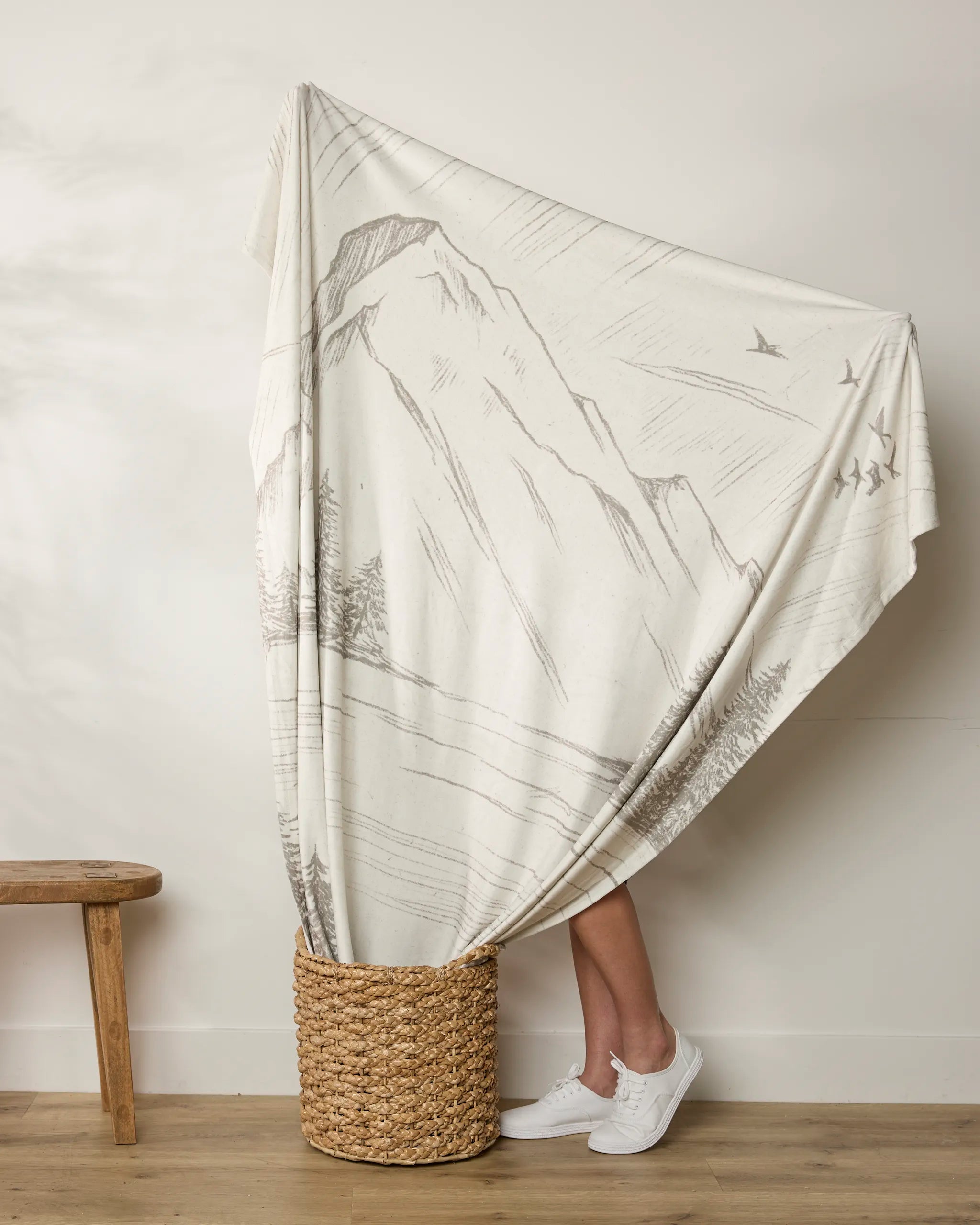 A person stands in a woven basket, wrapped in the Geometry "Stillpine" butter blanket, featuring mountain and bird designs. Only their lower legs and white sneakers are visible. A wooden bench is set against a plain wall nearby.