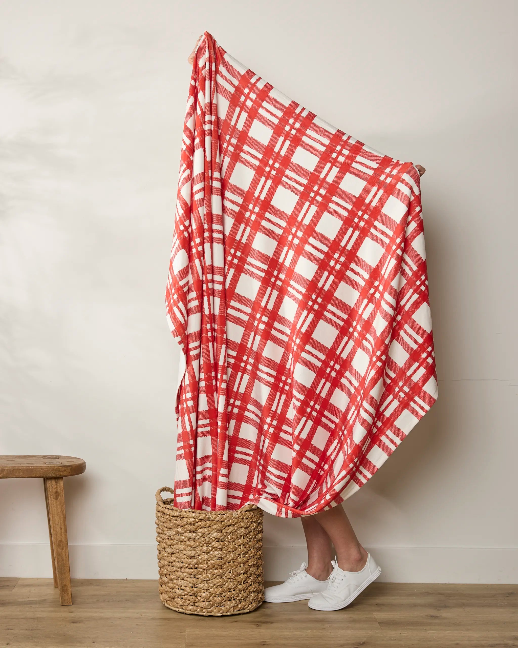 A person stands in a woven basket, wrapped in Geometry’s Tartan blanket, with only their lower legs and white sneakers visible. A wooden bench sits to the left against a white wall.
