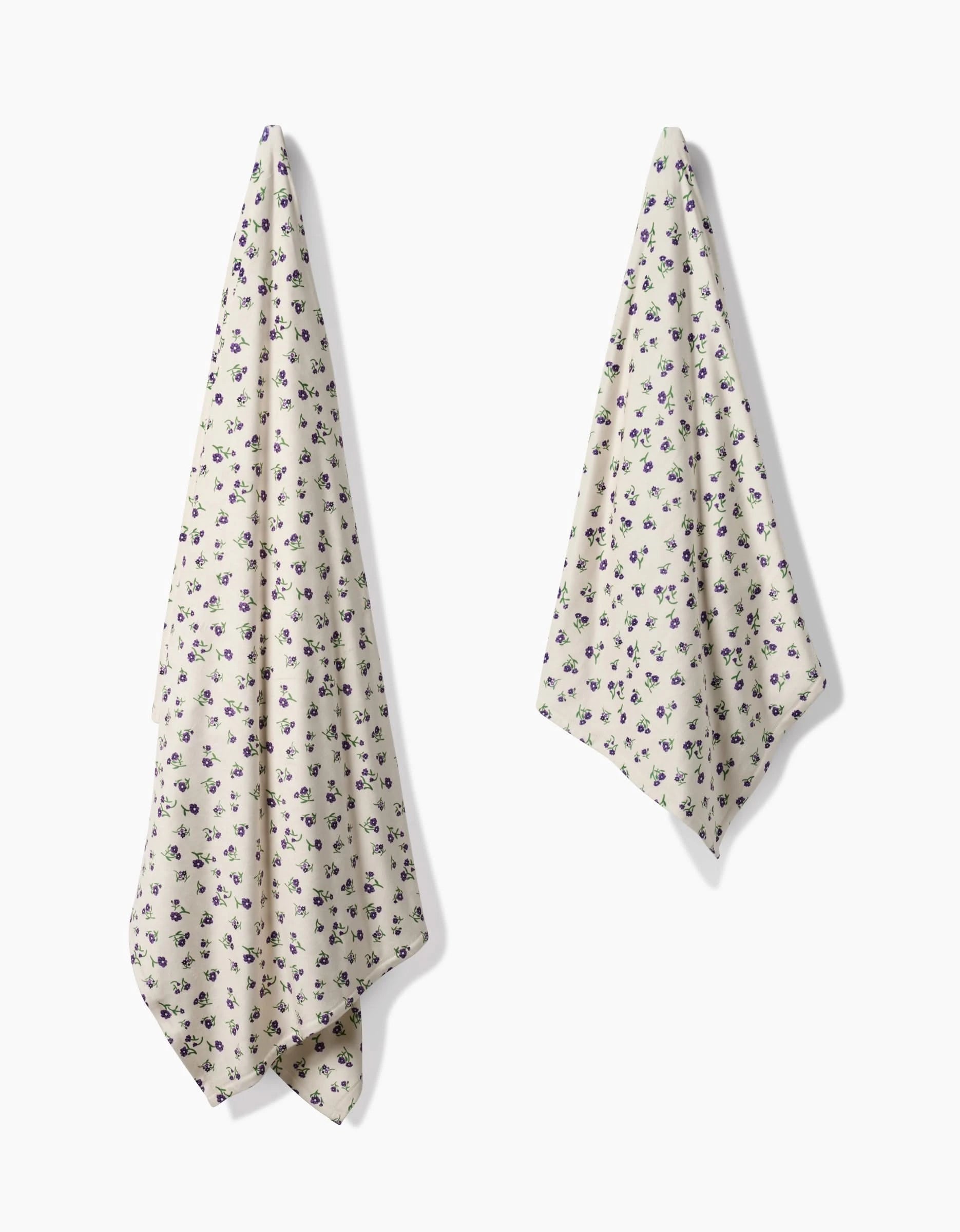 Two cream-colored towels with a small purple floral pattern hang on a white wall, offering the same soft elegance as Geometrys Violet Sound towels. One is larger and hangs loosely; the other is smaller with a sharper fold.