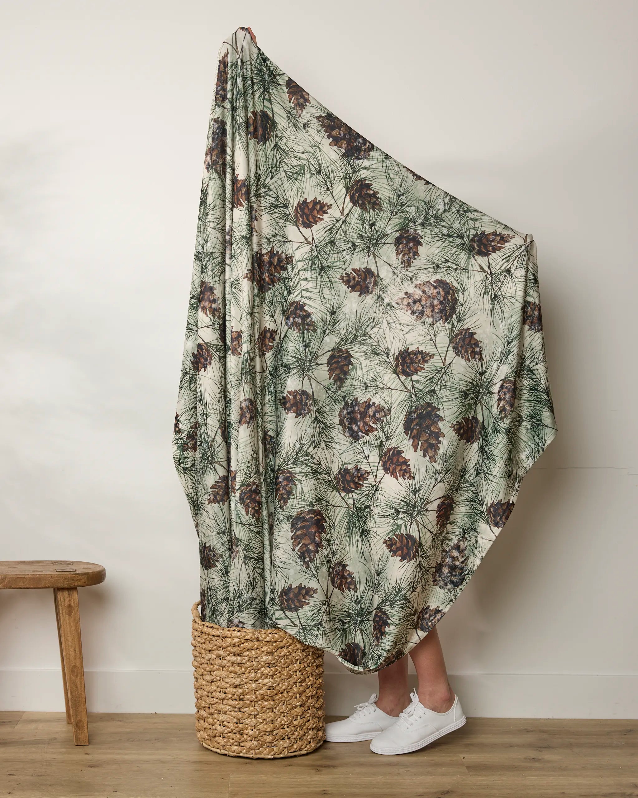 A person stands in a woven basket, mostly covered by Geometry’s Winter Pine Camo throw, featuring pine cone and branch patterns. Only their lower legs and white sneakers are visible beside a wooden bench and a plain wall.