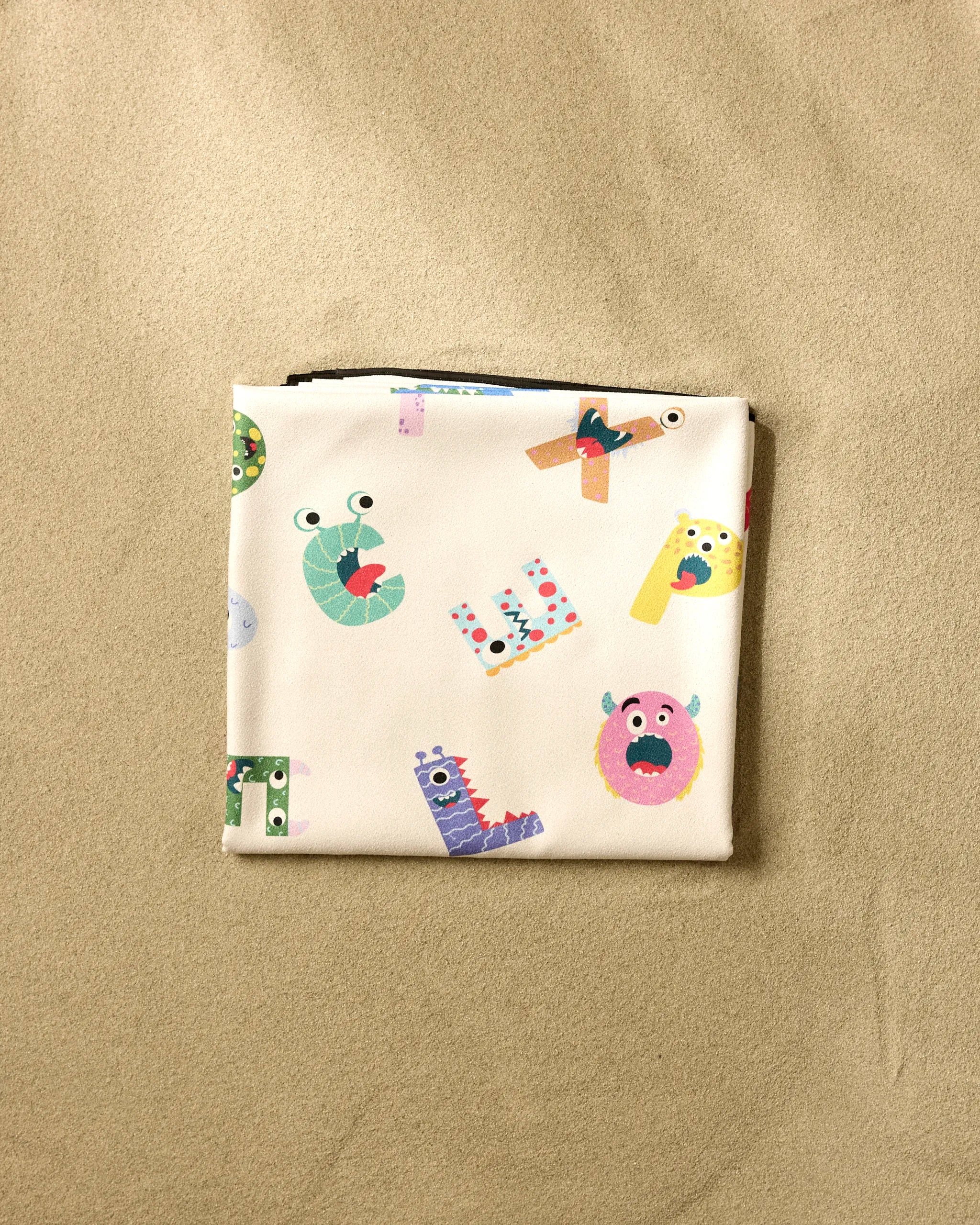 The Vaishali Design Studio "Alphabet Monsters" beach blanket features colorful cartoon letters and monster faces on a light background, displayed folded on a beige textured surface.