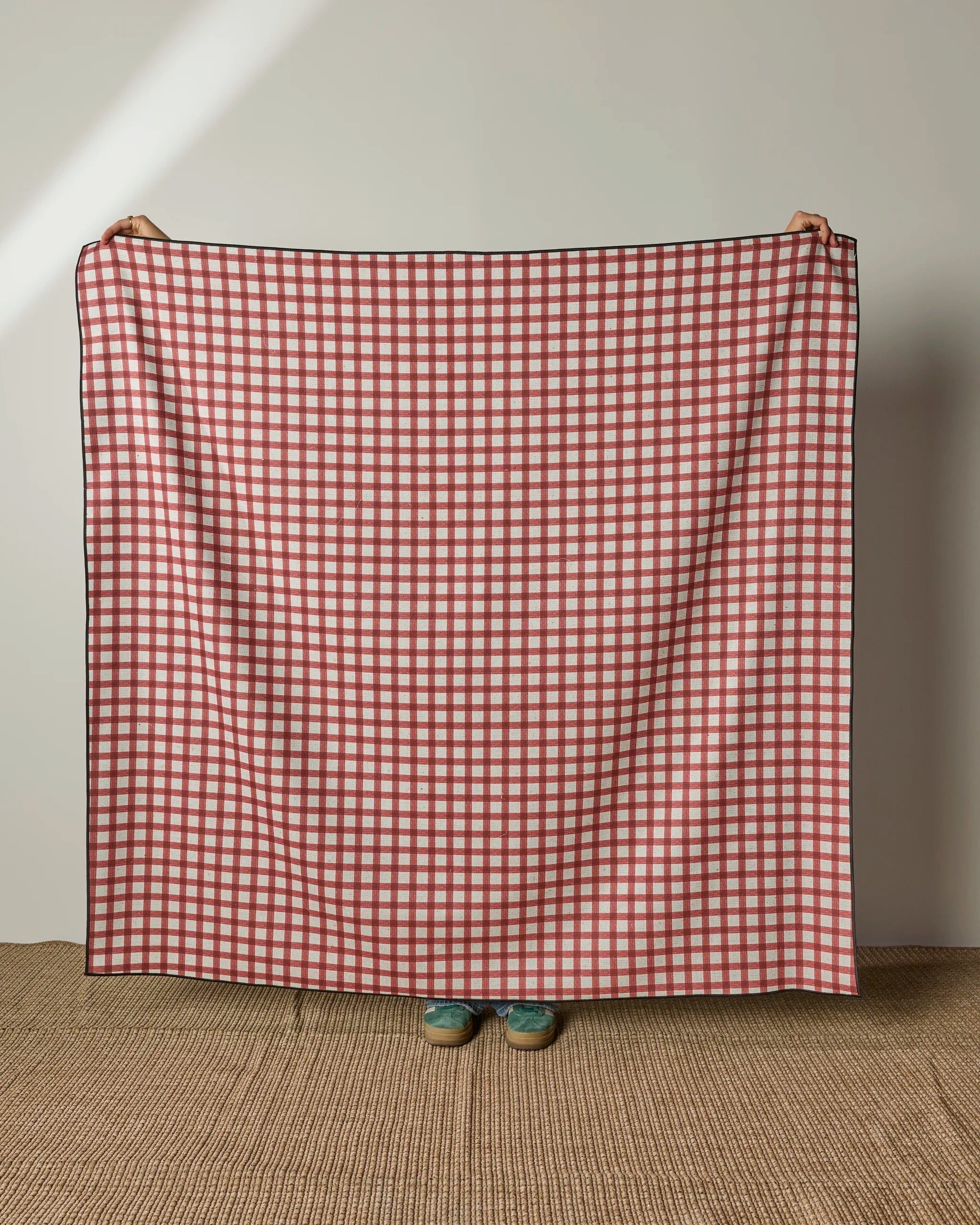 A person stands on a woven rug, holding up Geometry’s Apple Picnic beach blanket—a large red and white checked throw made from quick-drying, recycled materials—with only their hands and blue shoes visible behind it.