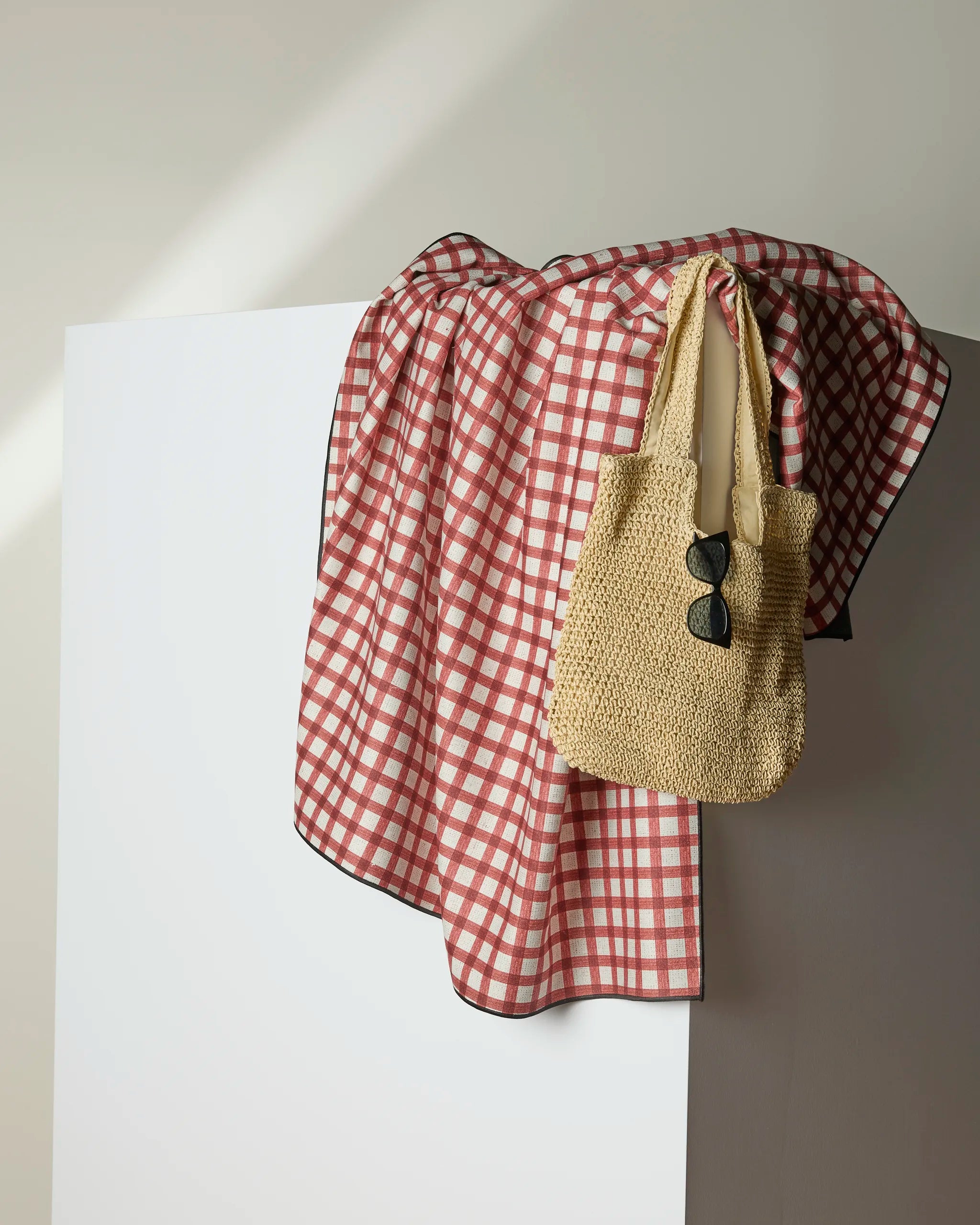 The Geometry Apple Picnic blanket in a red and white checkered pattern covers a white rectangular surface, topped with a recycled woven straw tote bag and black sunglasses hanging from the handle.