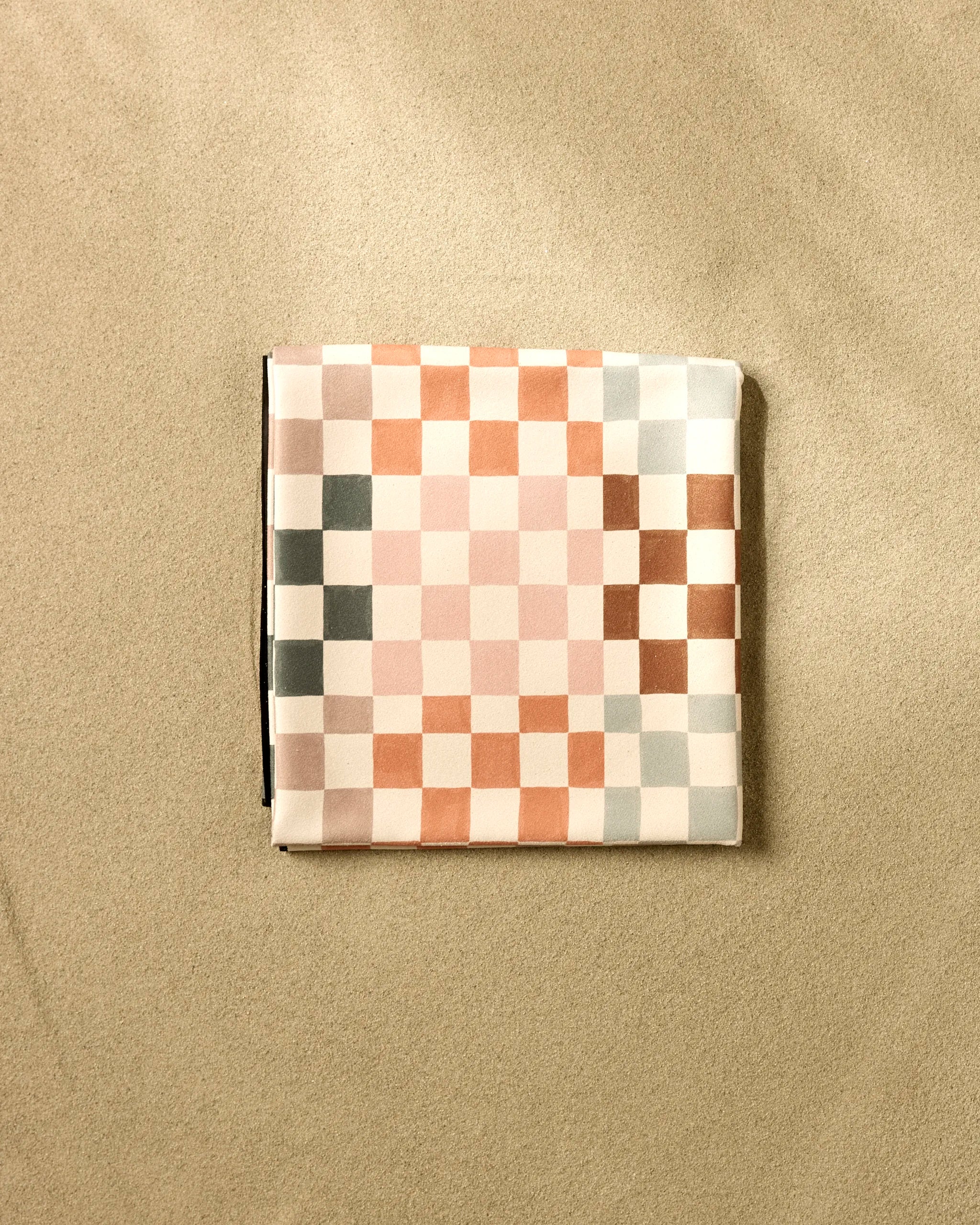 The GEOMETRY Autumn Checkers folded recycled beach blanket, featuring a muted pink, brown, gray, and white checkerboard pattern, rests on a sandy beige surface.