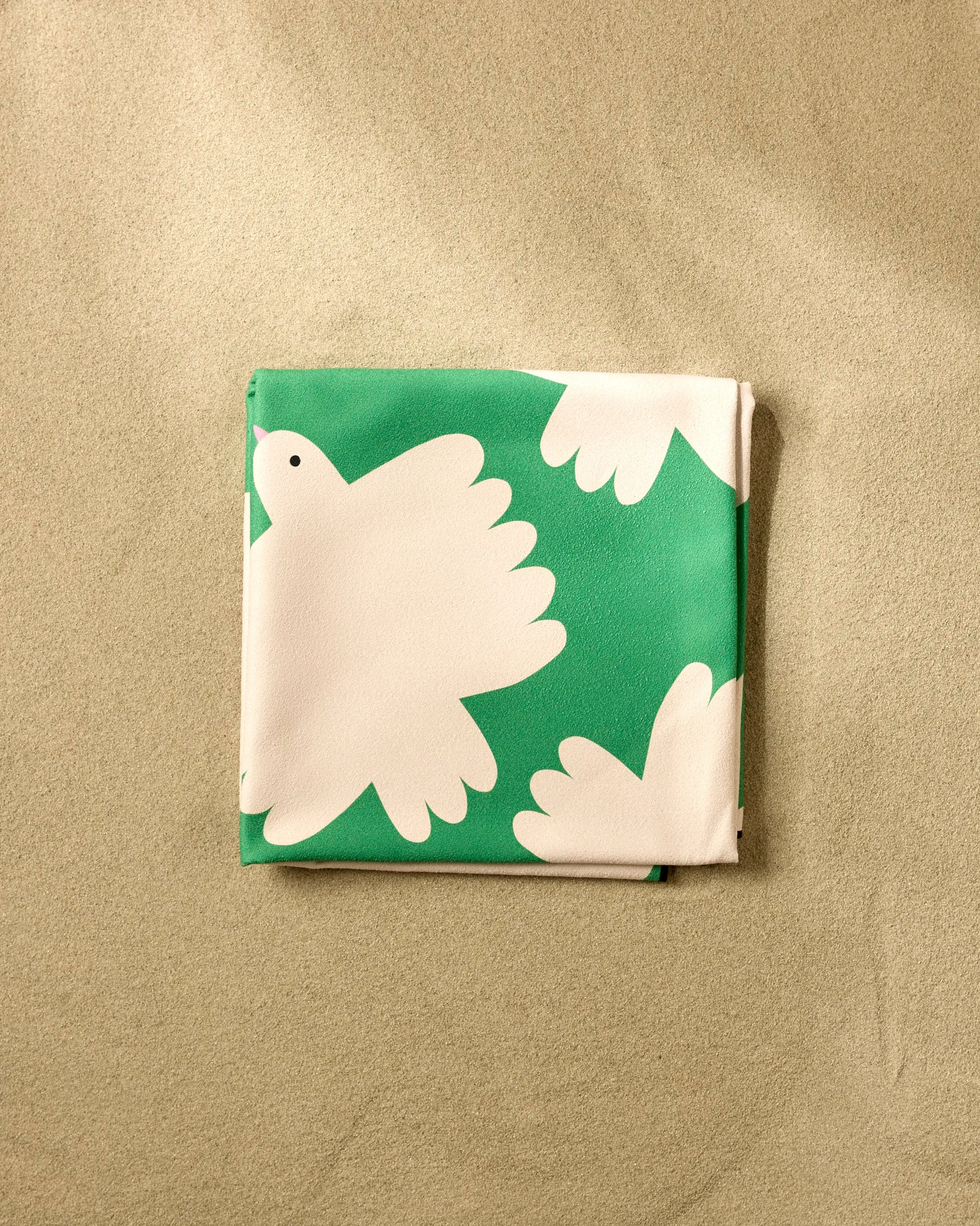 A neatly folded green "Birds" beach blanket by Jennifer Bouron, featuring white dove patterns and made from recycled materials, rests on a sandy beige surface.