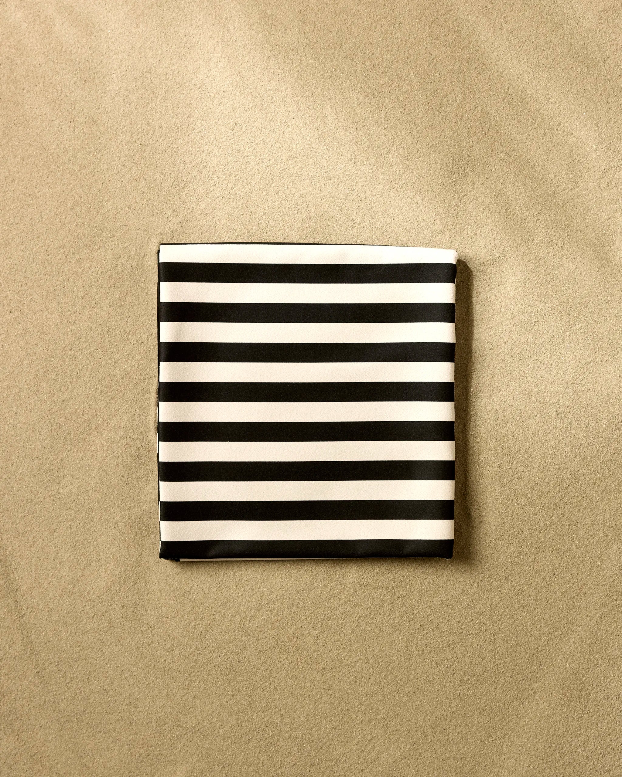 The GEOMETRY Black Stripes beach blanket, featuring bold black and white horizontal stripes, adds modern flair to the sandy beach.